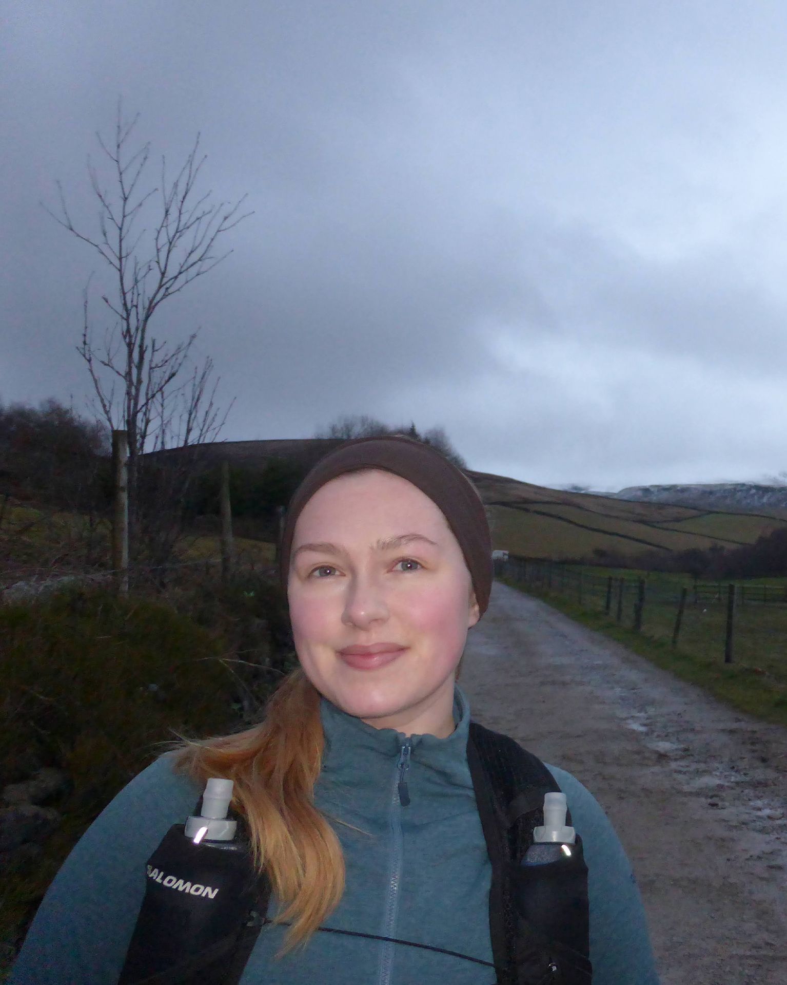 Person wearing outdoor sports gear on a rural path with hills and a cloudy sky in the background.