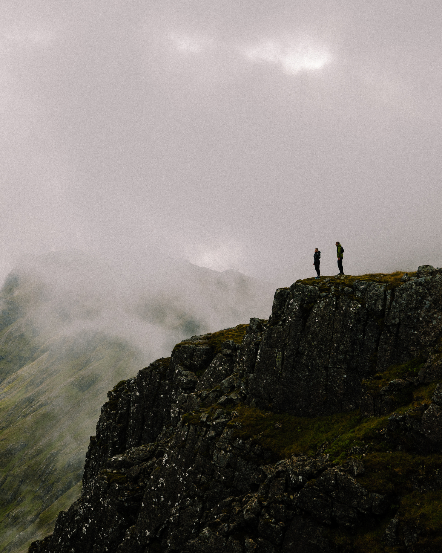 Two hikers stand on a rugged cliff edge, surrounded by misty mountains and a cloudy sky.