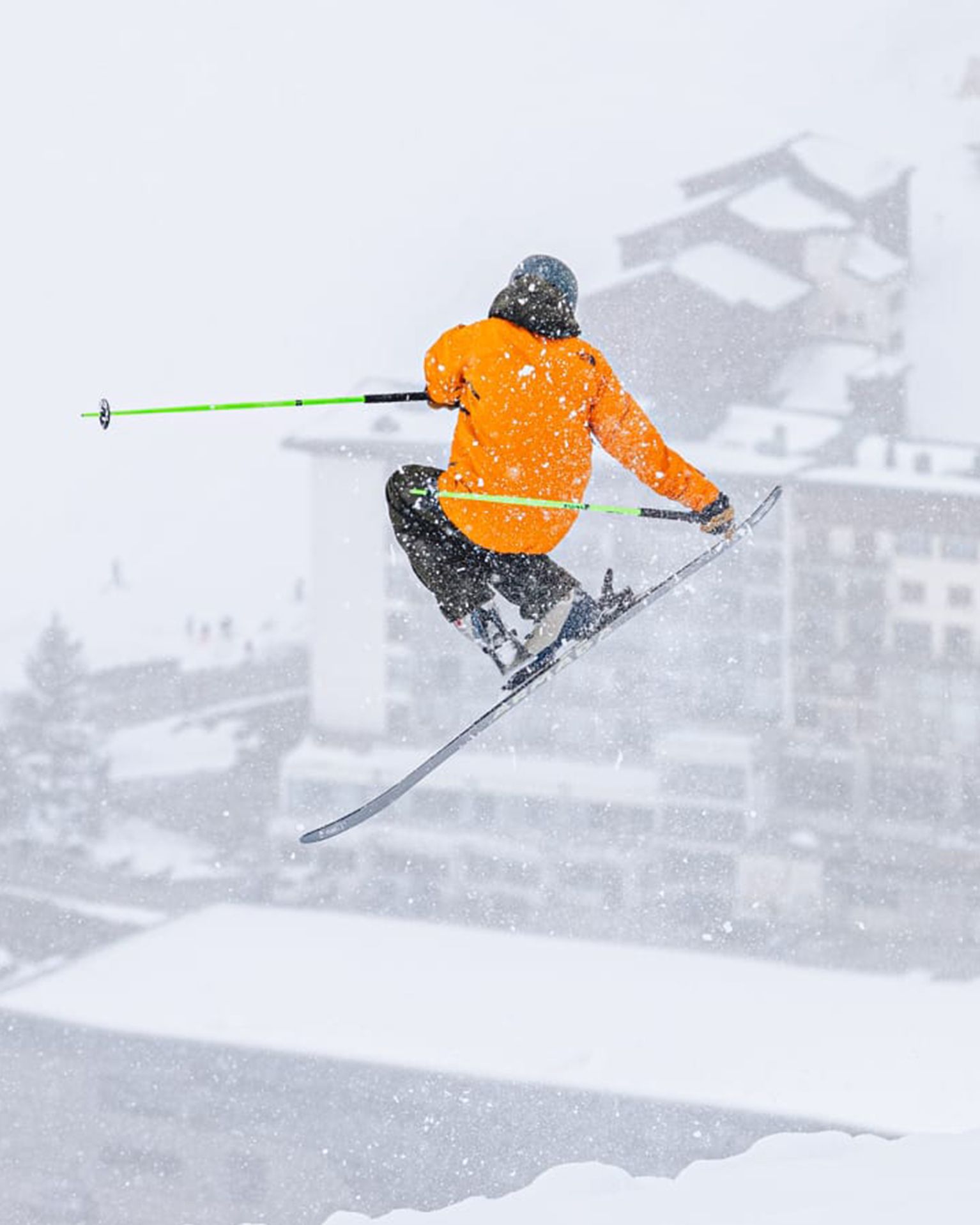 Skier in an orange jacket mid-air, performing a jump on snowy mountain backdrop with a blurred view of buildings below.