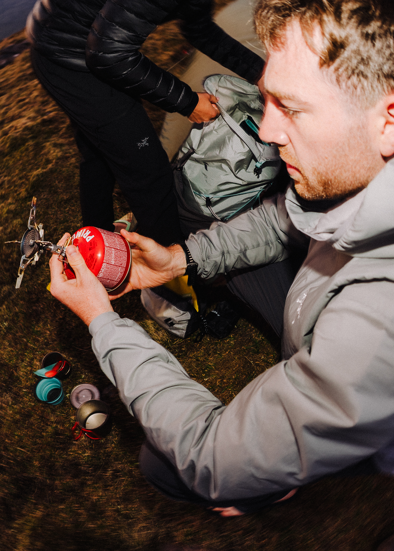 Man assembling a portable camping stove outdoors, with various cooking gear on the ground nearby.
