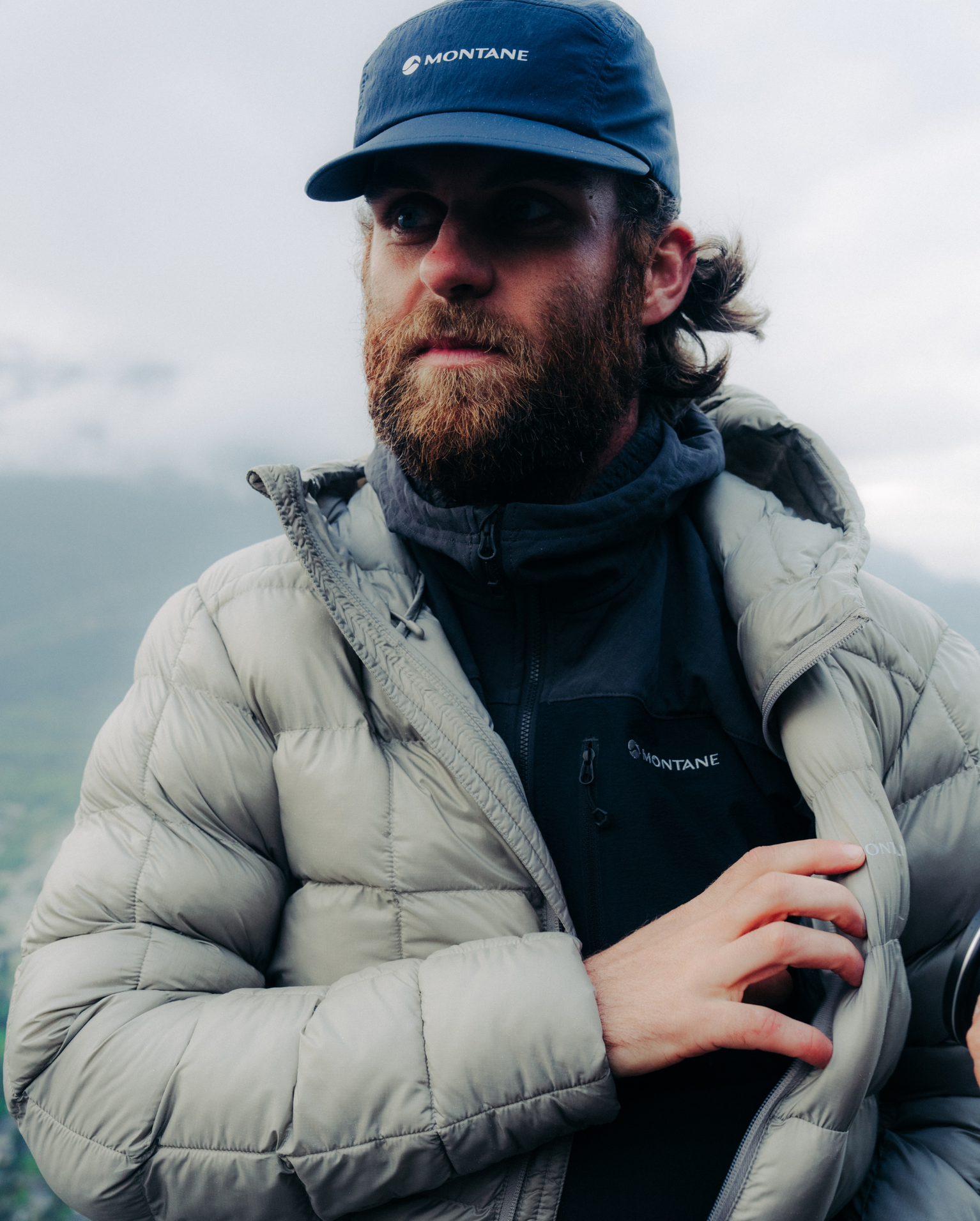 A man wearing a grey puffer jacket and blue cap, standing outdoors with mountains in the background.