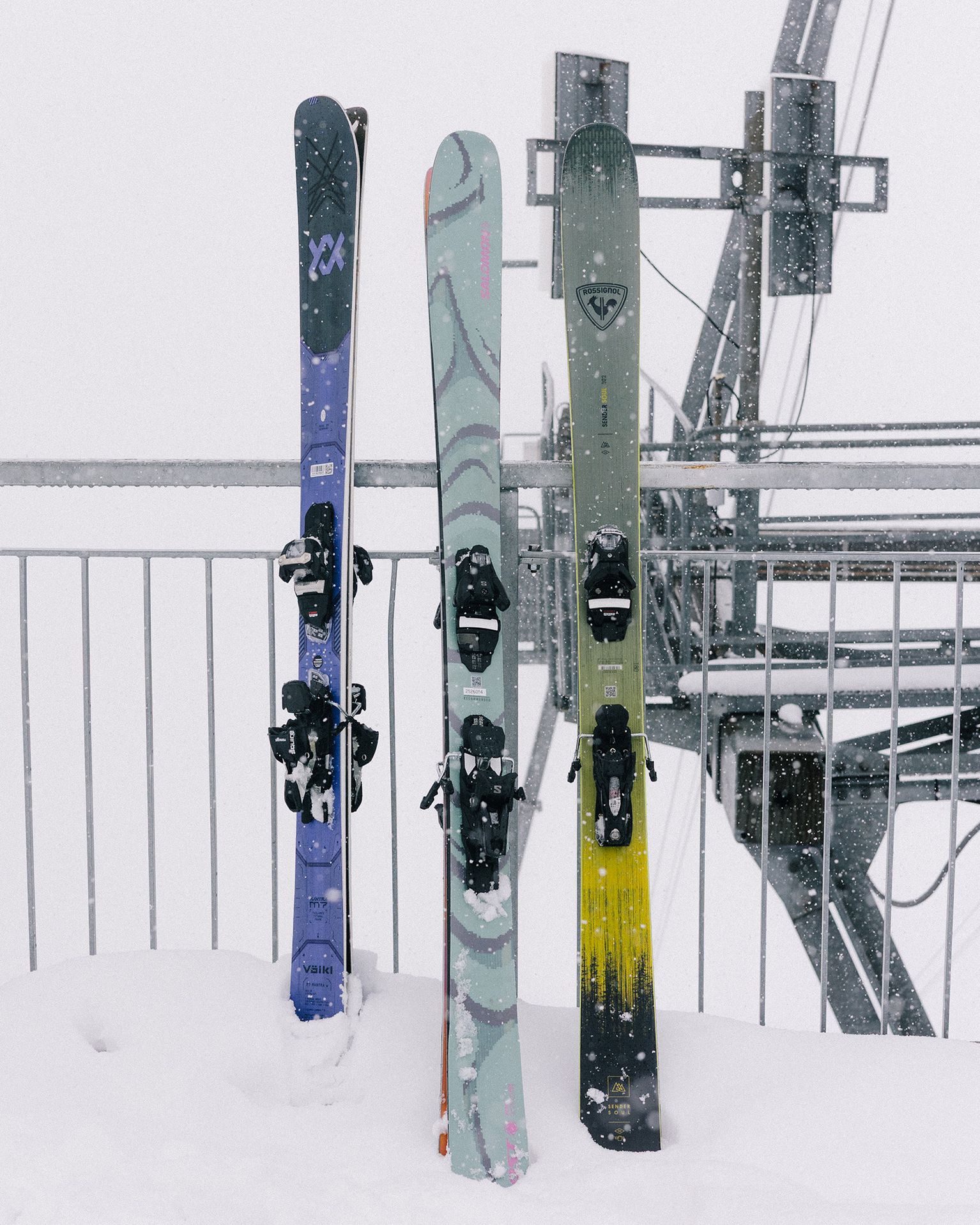 Three pairs of skis standing upright in the snow, with a snowy backdrop and ski lift machinery visible in the background.
