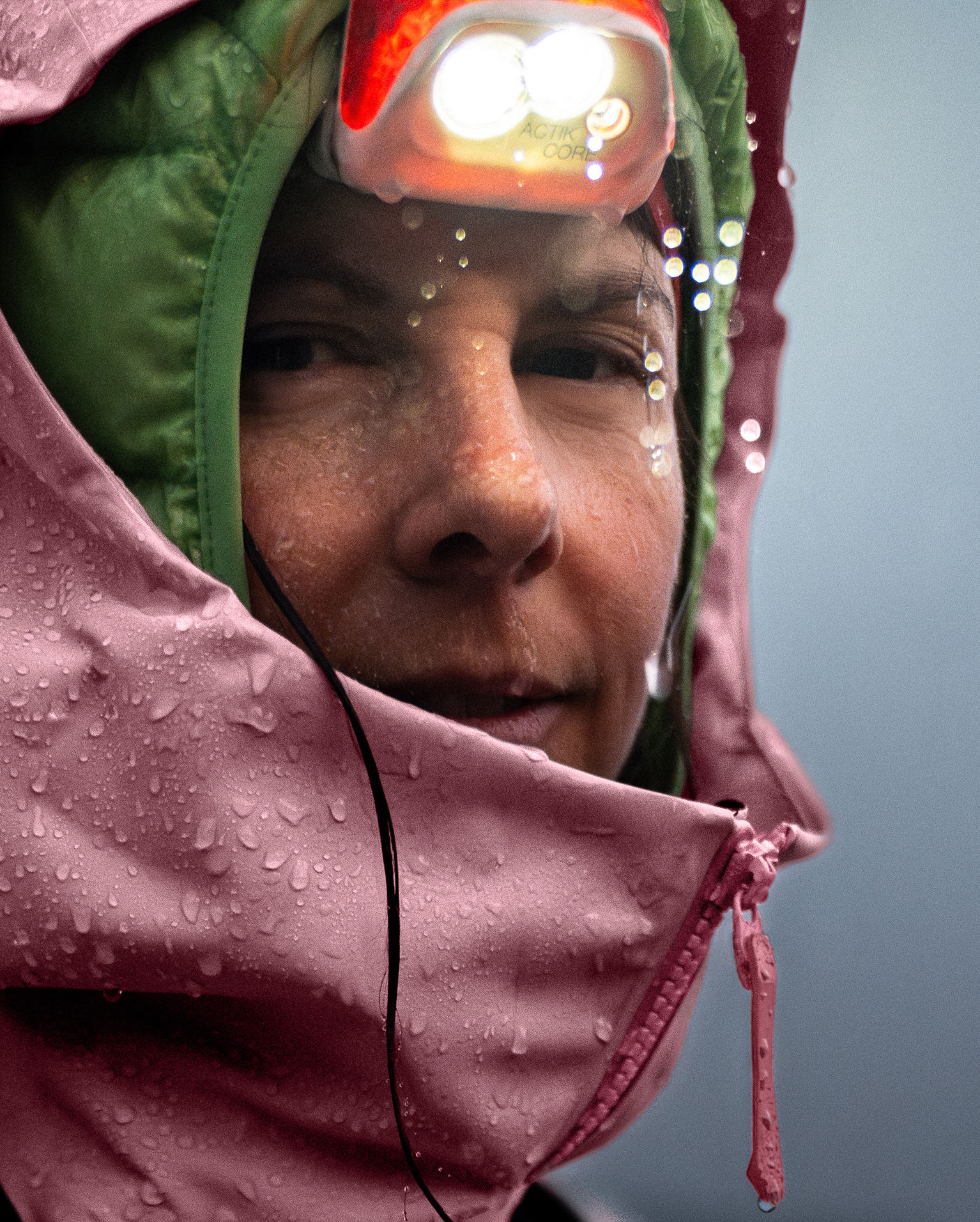 Woman wearing an Arc'teryx Beta SV jacket and a headlamp, standing in the rain. Water droplets cover the jacket, emphasising rain and cool conditions.