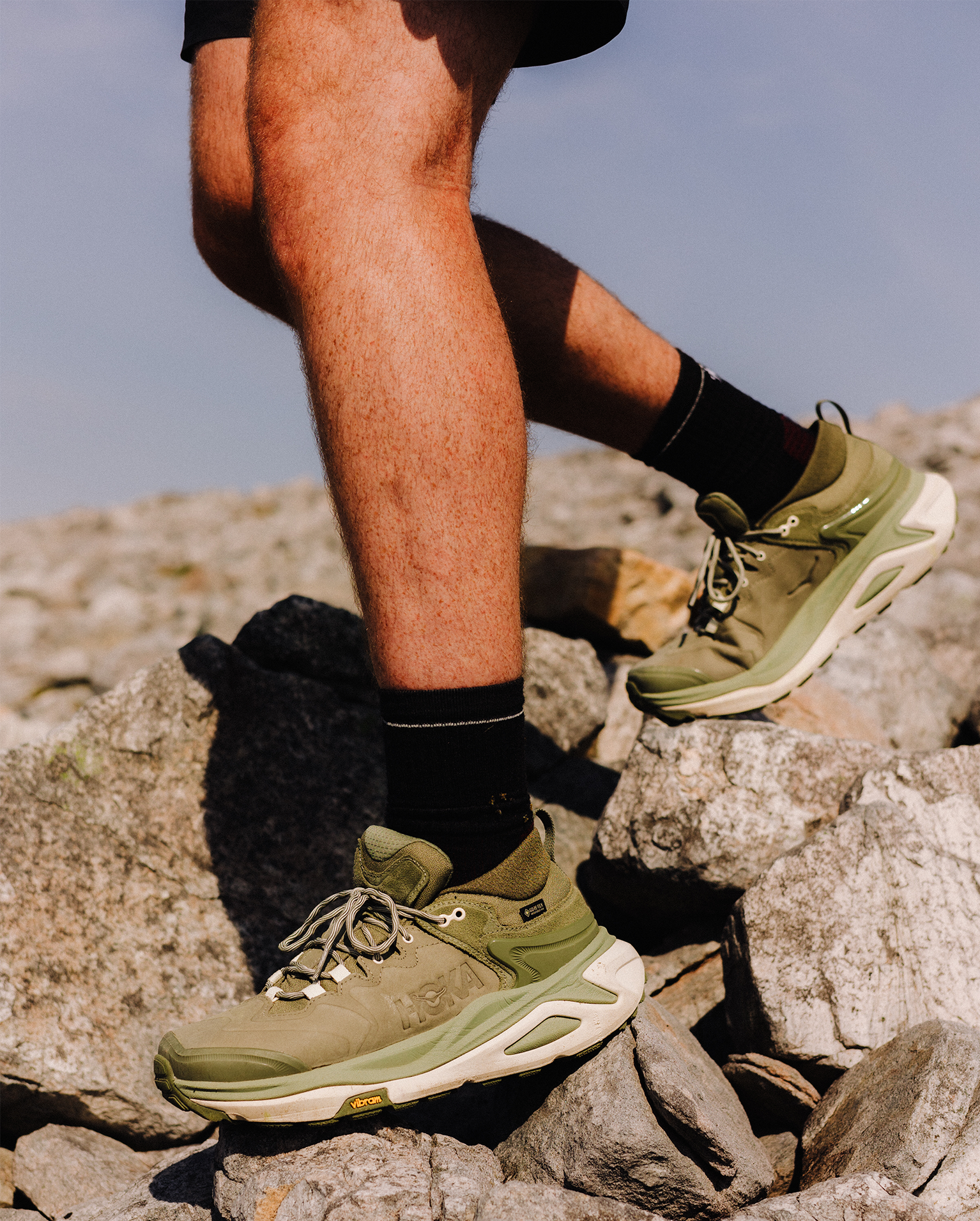 Close-up of a climber wearing green hiking shoes and black socks, ascending a rocky terrain under a clear sky.
