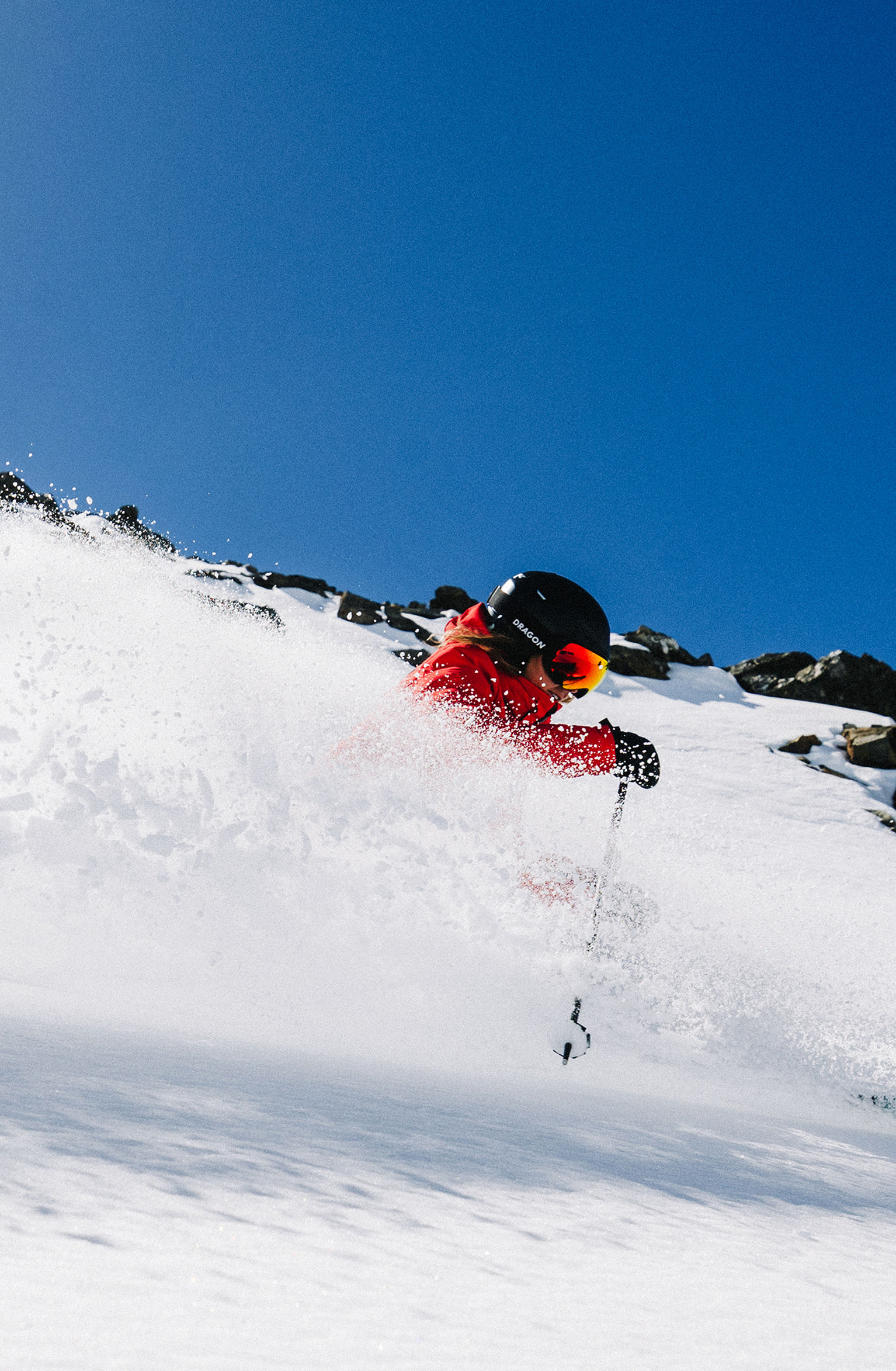 Skier in a red outfit glides swiftly down a snowy mountain slope under a clear blue sky, leaving a trail of powdery snow behind.