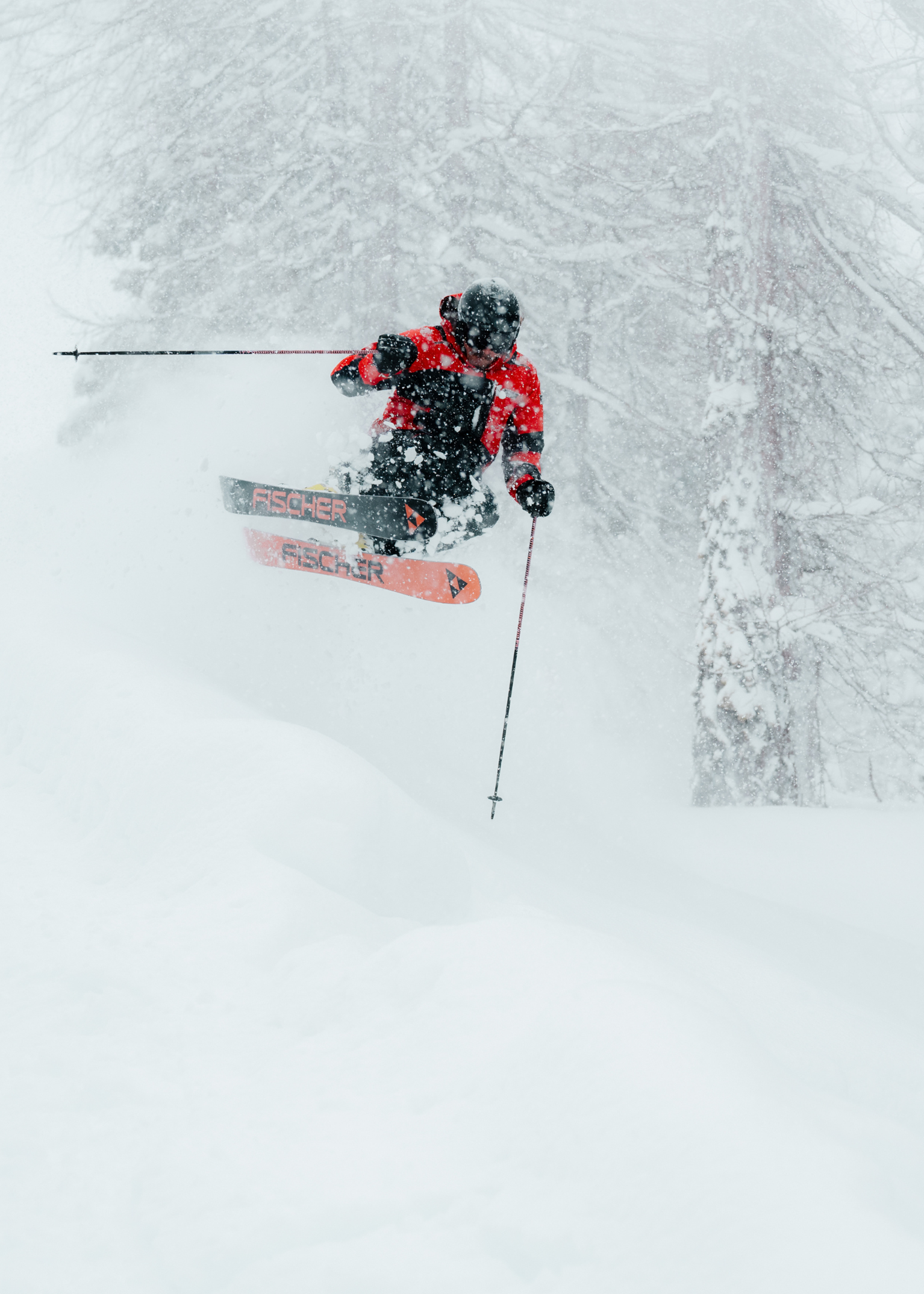 Skier in red jacket and black helmet launching through deep powder by snow-laden trees, orange Fischer skis visible.