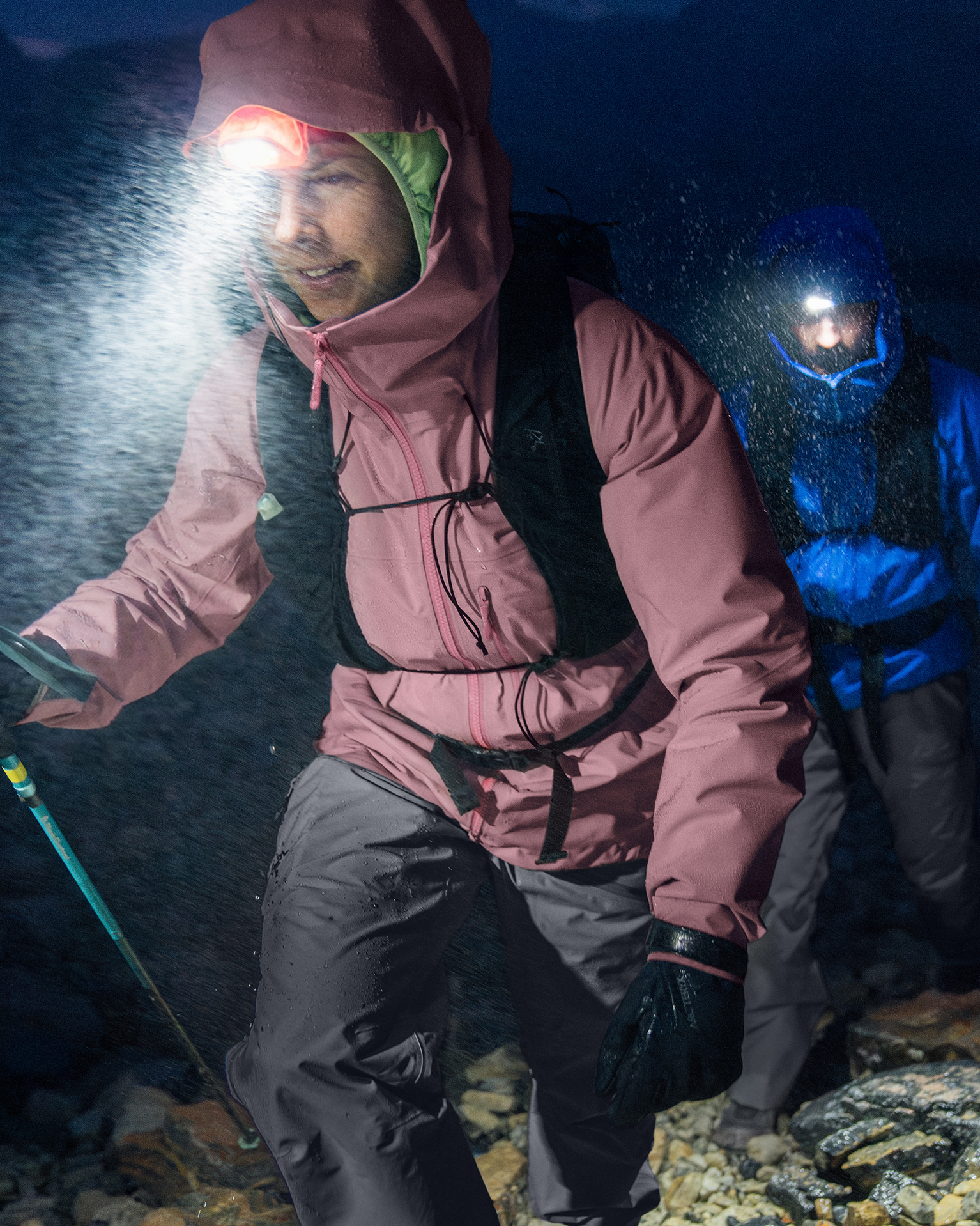 Two hikers in waterproof gear trek through rain at night, headlamps illuminated, on a rocky mountain path.