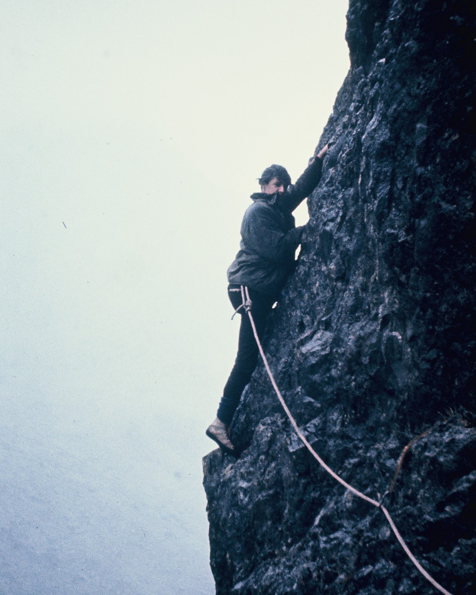 A climber ascends a steep, rocky cliff face, secured by a rope, against a foggy sky backdrop.