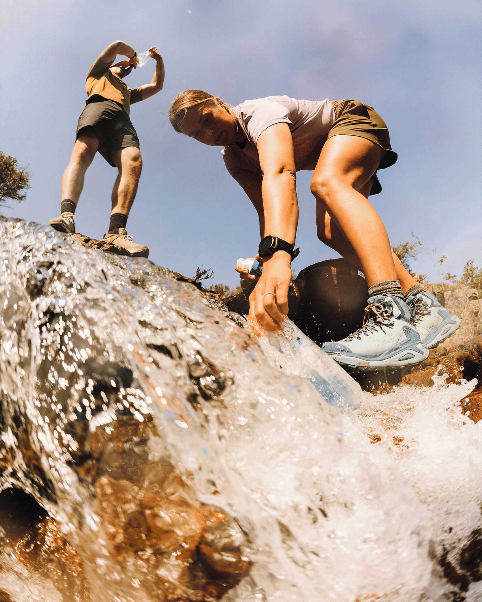 Two hikers by a stream, one filling a bottle with water in a mountain setting.