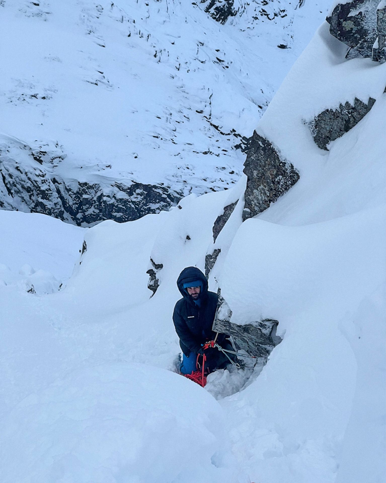 A climber wearing winter gear ascends a snow-covered, rocky slope, using ropes for support in a rugged mountain environment.
