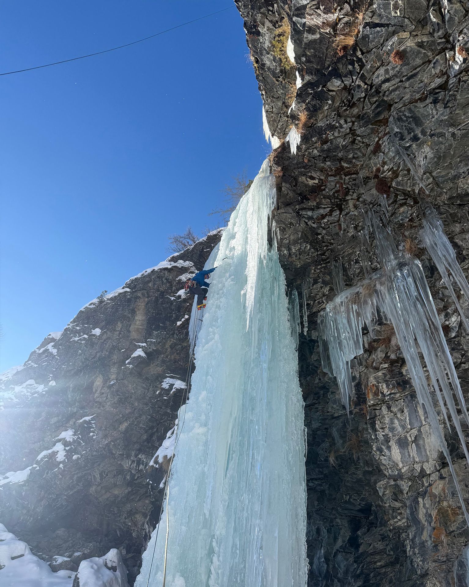 A climber ascends a vertical ice wall on a rocky cliff under a clear blue sky, surrounded by hanging icicles.