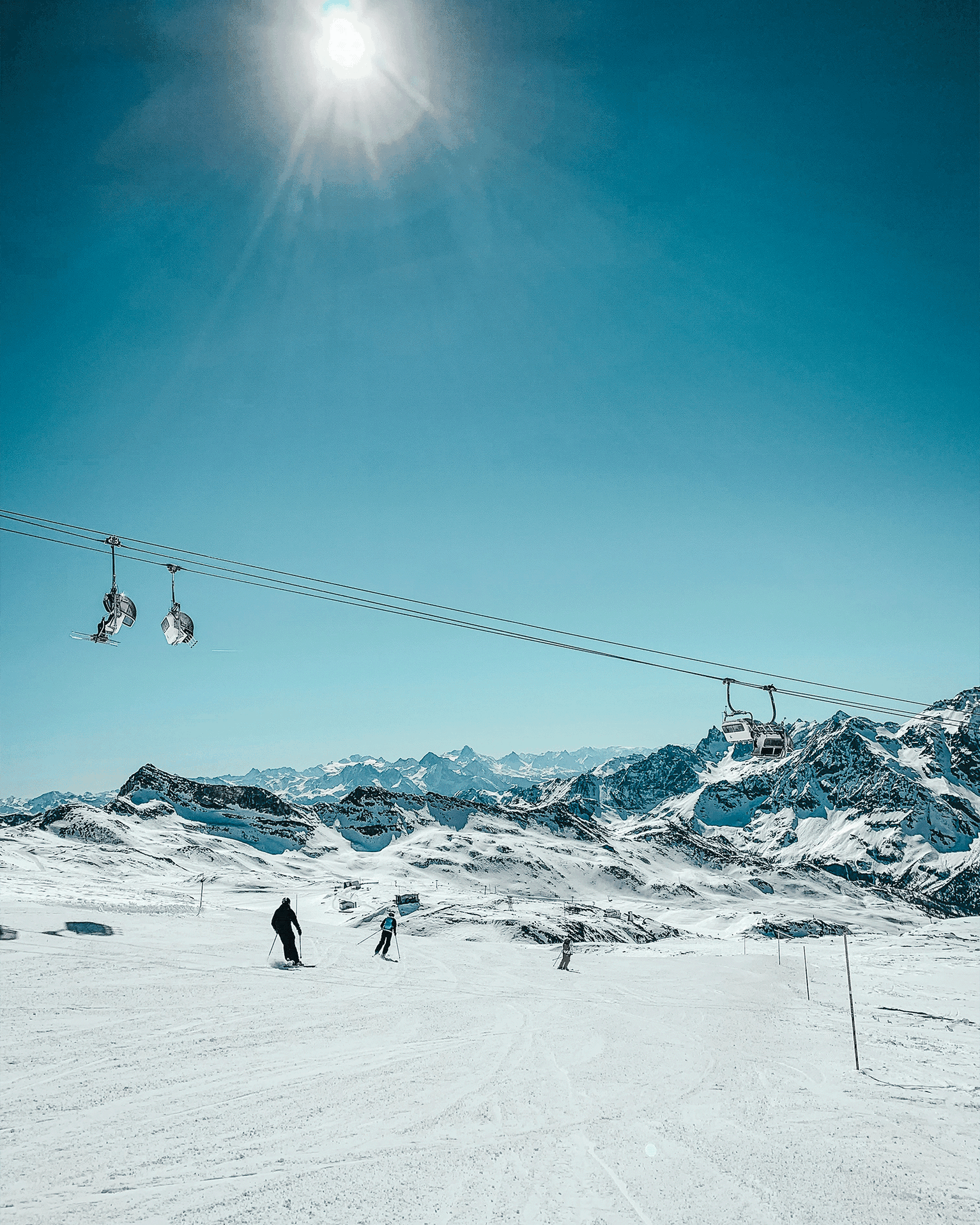 Skiers glide down a snowy mountain under a bright sun, with distant peaks and a ski lift in the background.