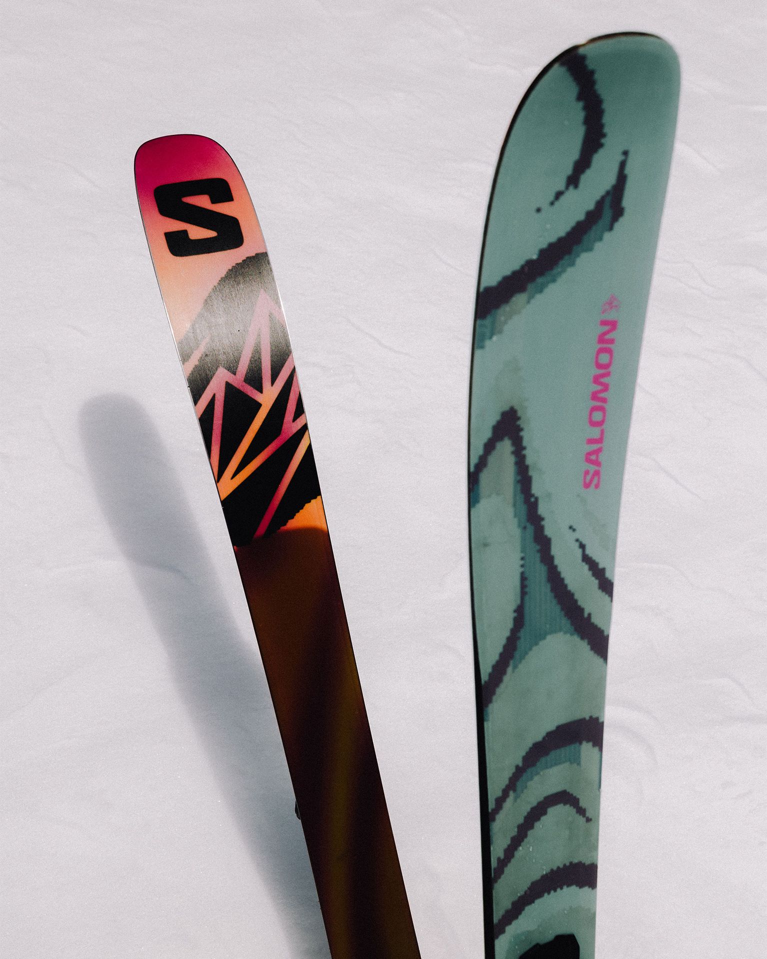Two skis standing upright in the snow, one with a colorful geometric design and the other with a green and black pattern, branded "Salomon."