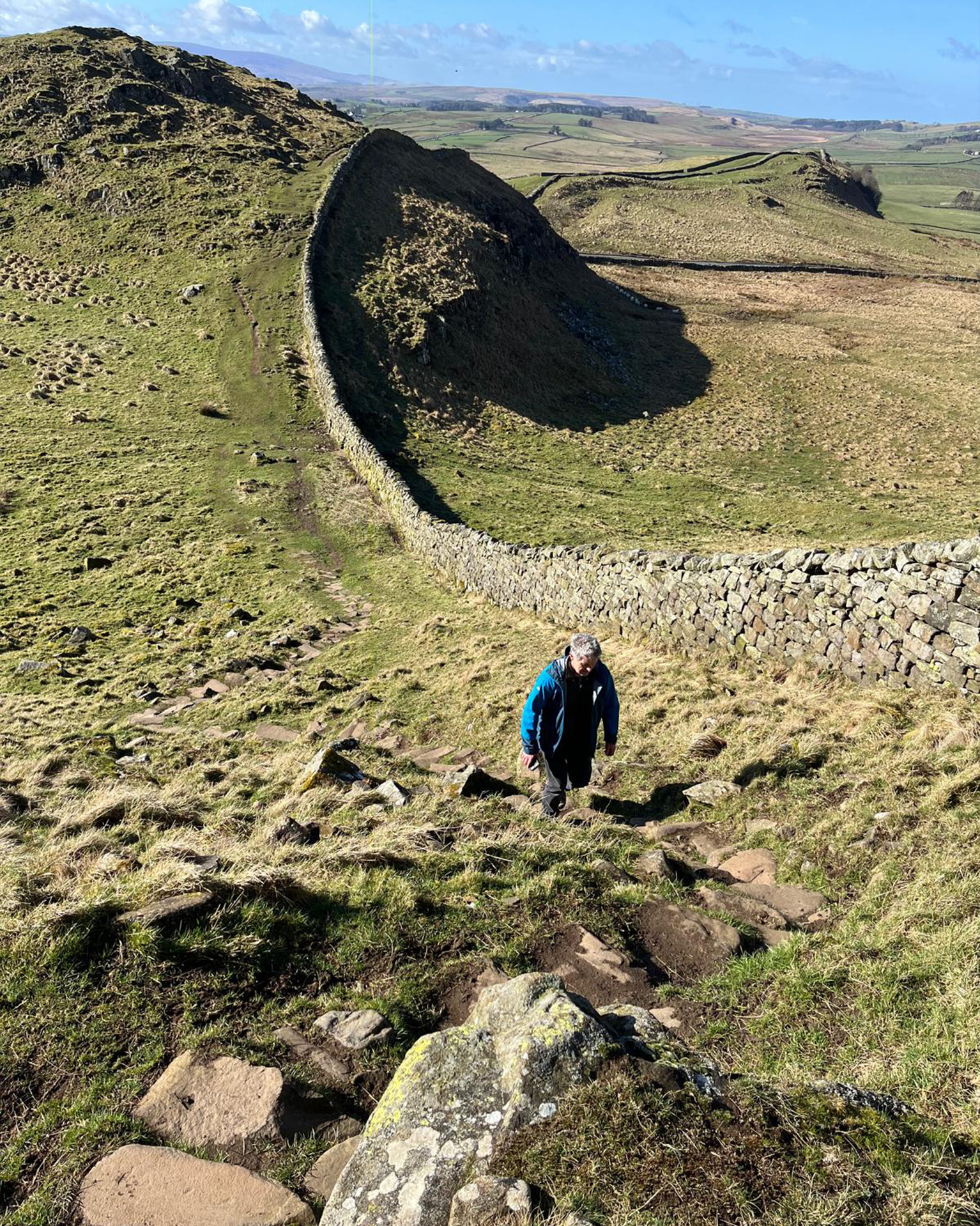 Person hiking on a grassy hilltop path with a stone wall, under a bright blue sky with scattered clouds.