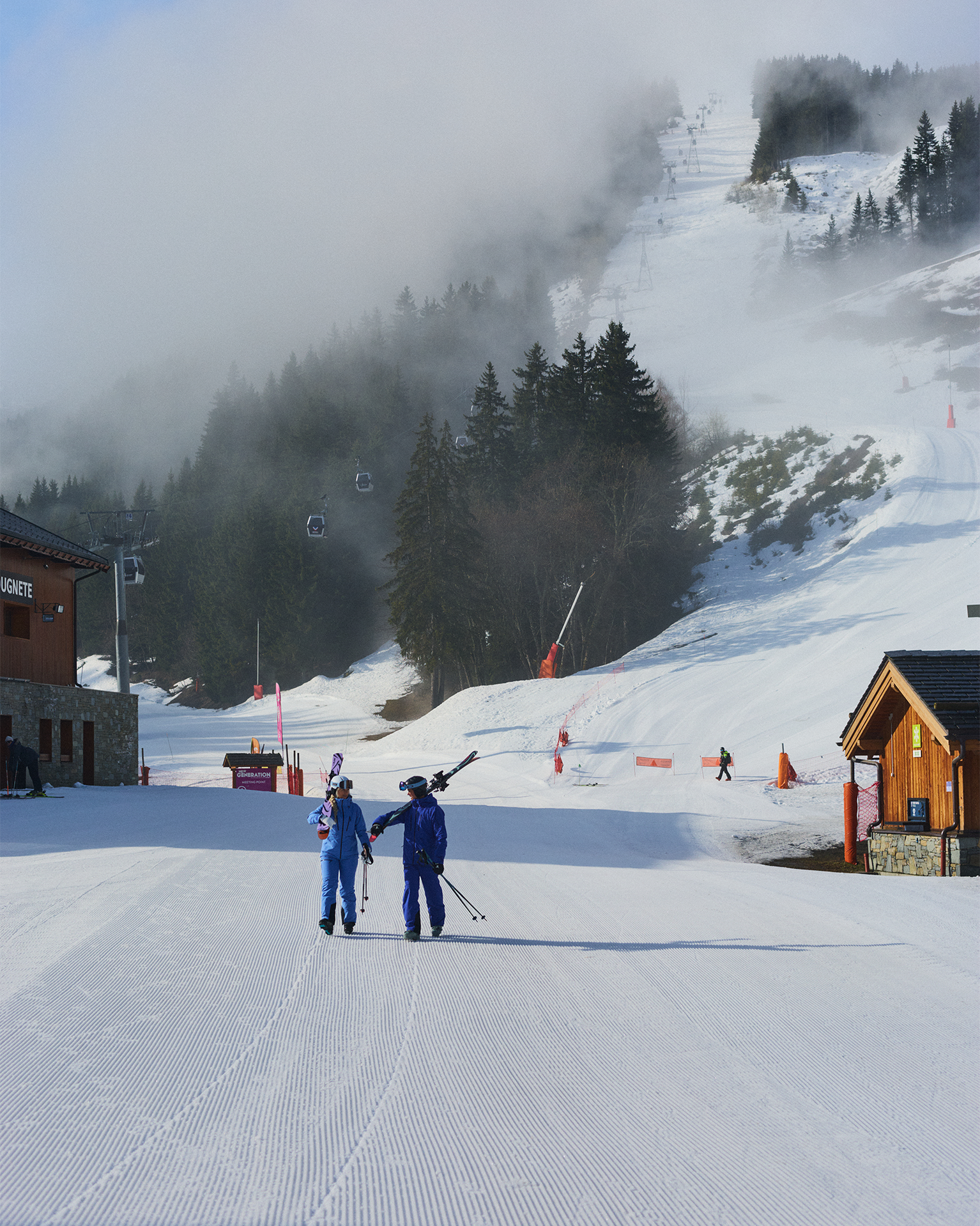 Two skiers in blue outfits walk up a snowy mountain slope, with fog rolling over pine trees and a ski lift in the background.