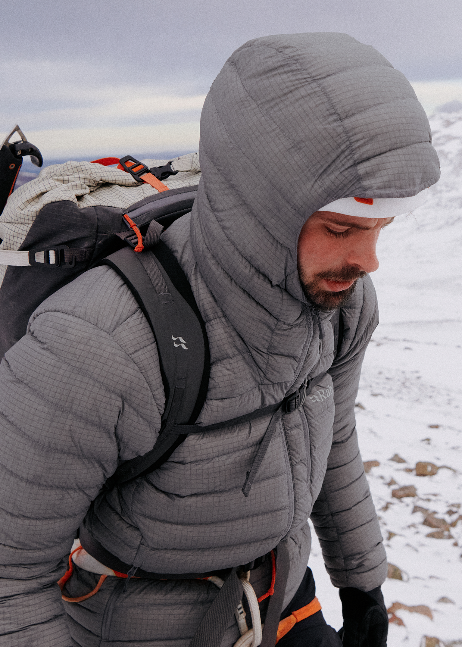 Mountaineer in grey insulated jacket and backpack walks through snowy terrain, focused and equipped for a challenging climb.