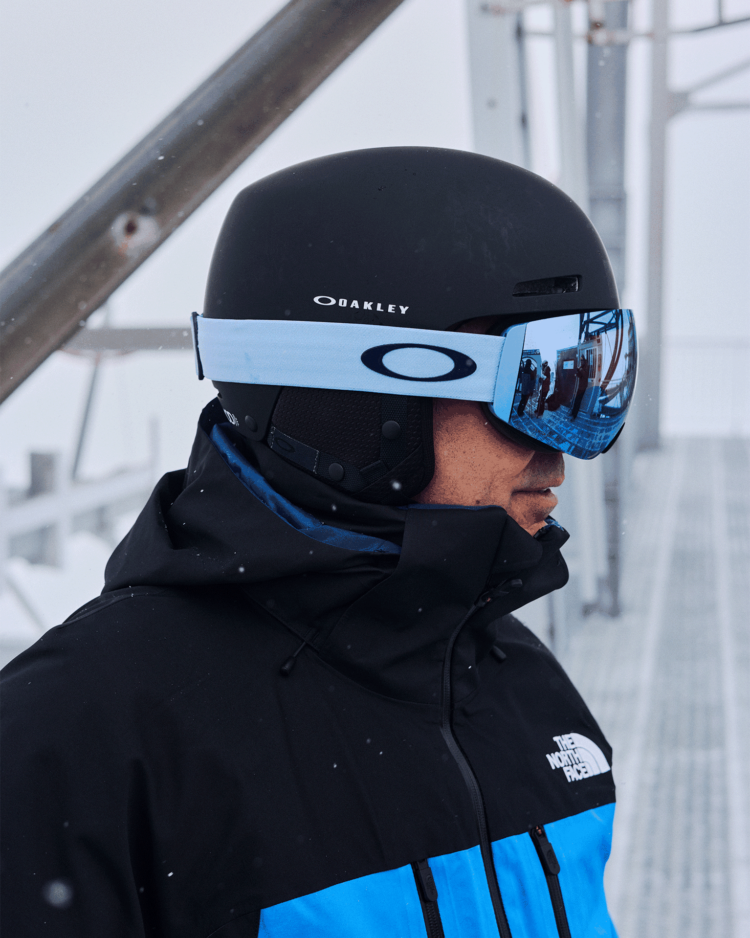 Man wearing a black helmet and blue-tinted goggles, dressed in a black and blue jacket with snowflakes falling around.