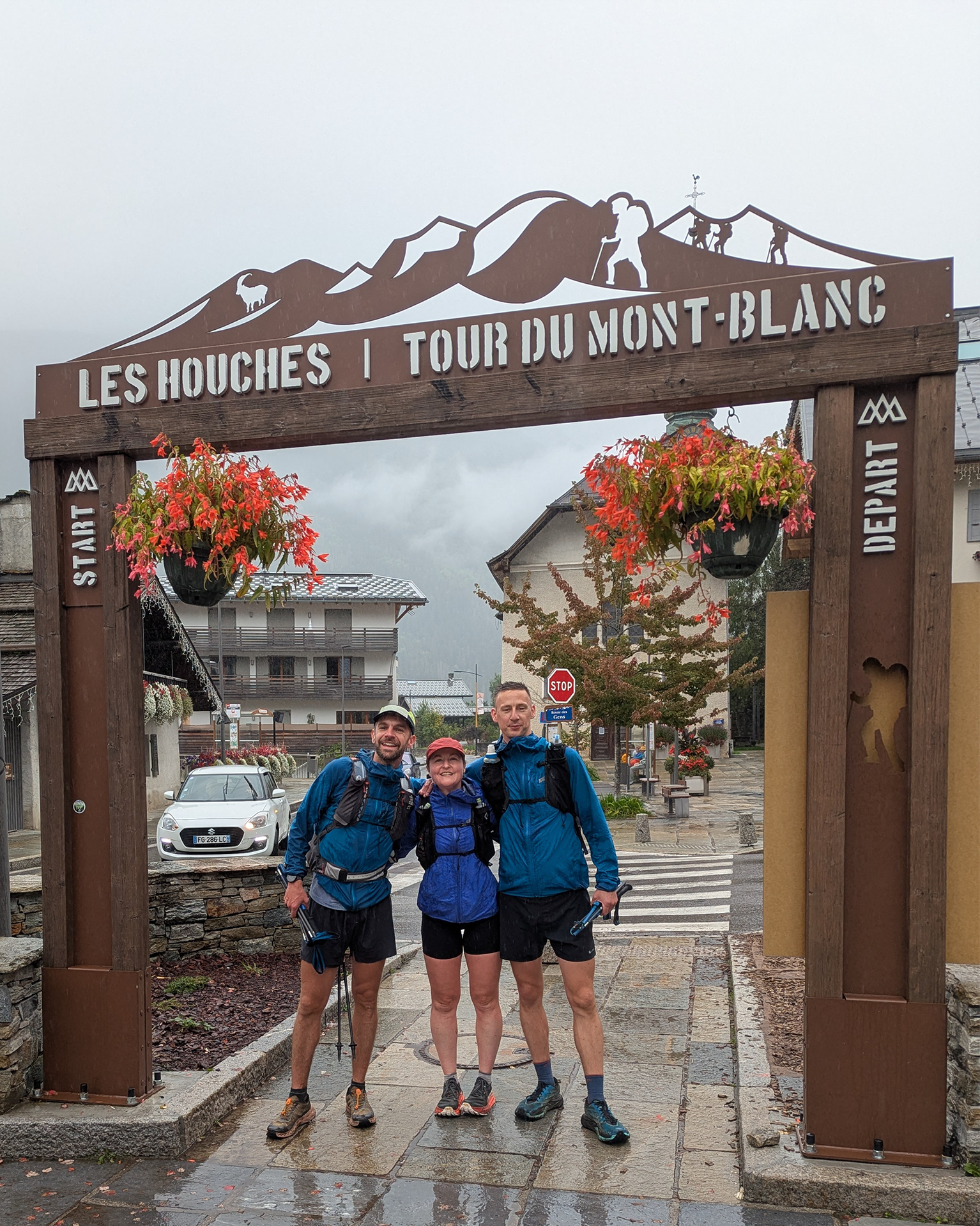 Three hikers in mountain gear stand smiling under the "Les Houches Tour du Mont-Blanc" archway, with misty mountains in the background.