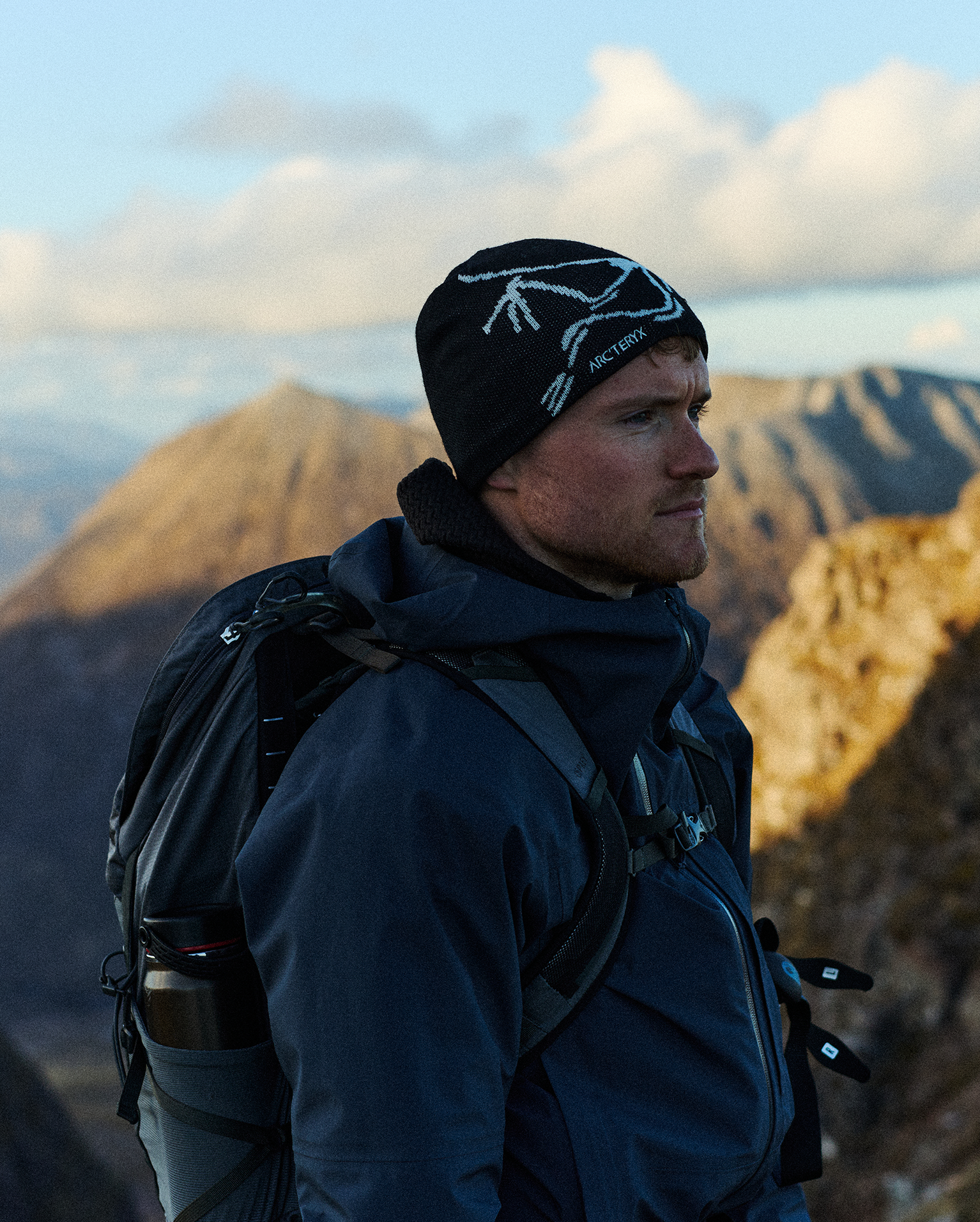 Man in navy outdoor gear and beanie, carrying a backpack, standing on a rocky mountain path with distant peaks and a cloudy sky in the background.