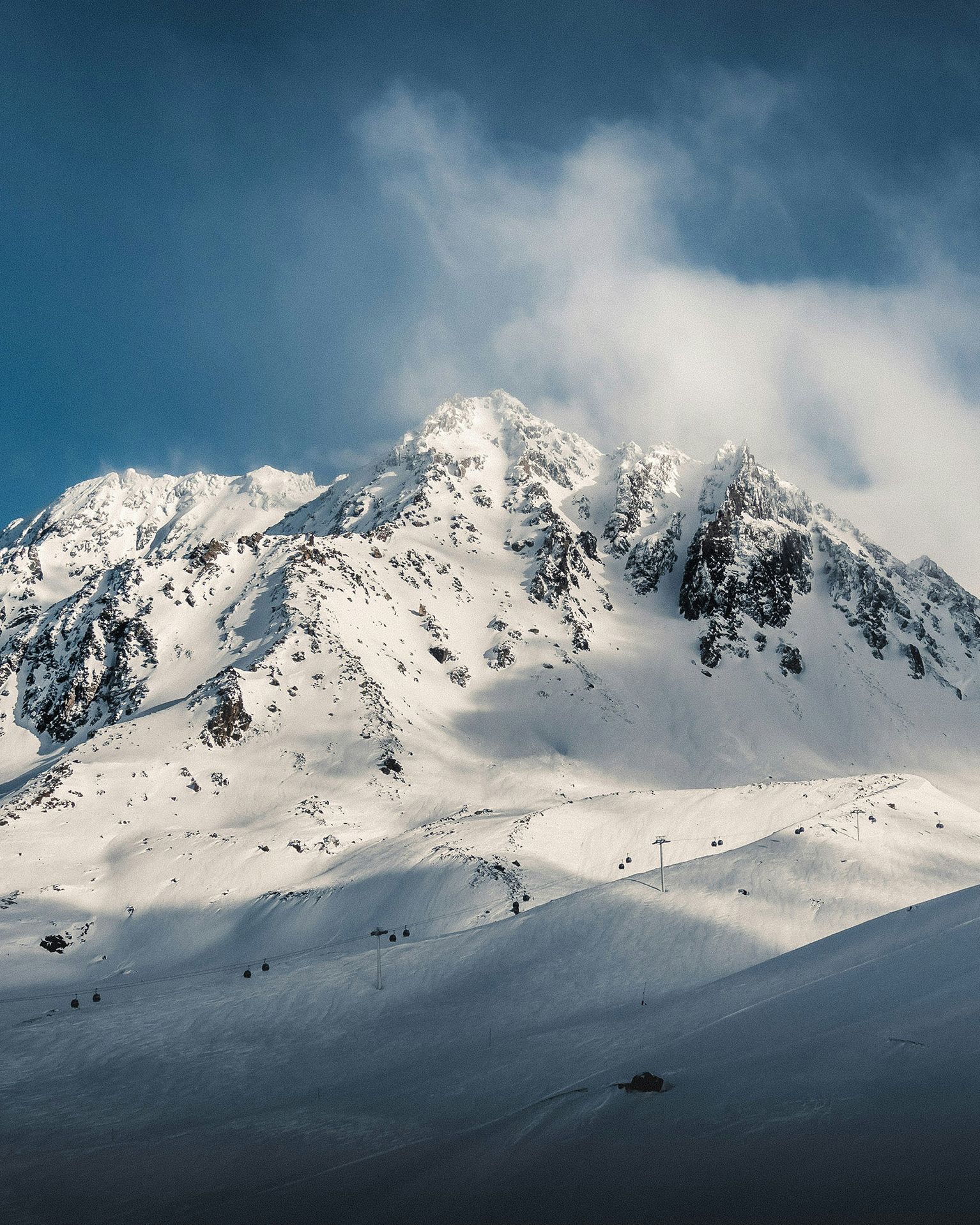 Snow-covered mountain under a clear blue sky, with rugged peaks and soft clouds drifting above, casting shadows on the slopes below.