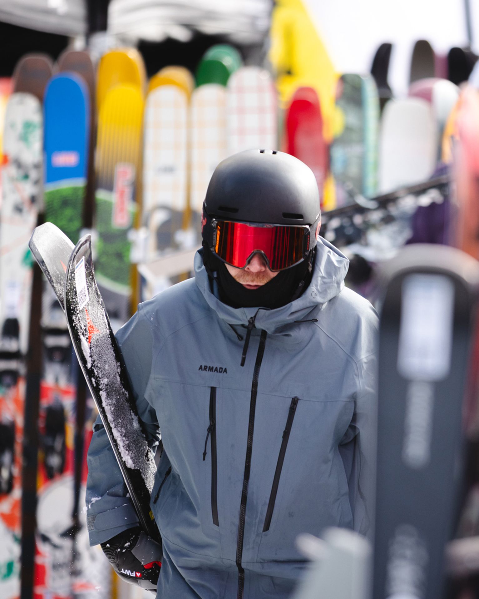 A person in a grey jacket and black helmet carries skis, surrounded by various colourful skis in a storage area.