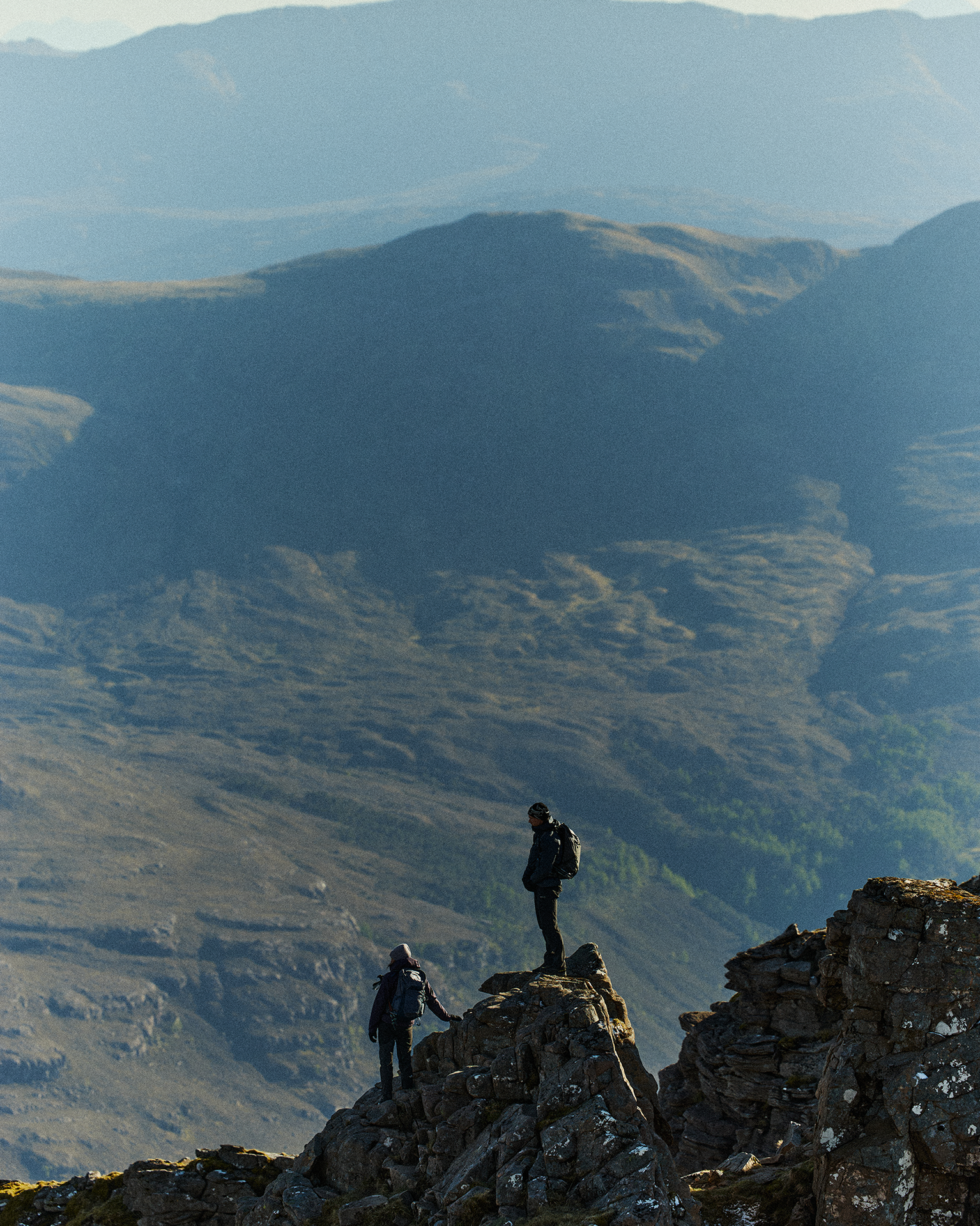 Two hikers in outdoor gear stand on rocky terrain, overlooking a vast mountain landscape under a clear sky.