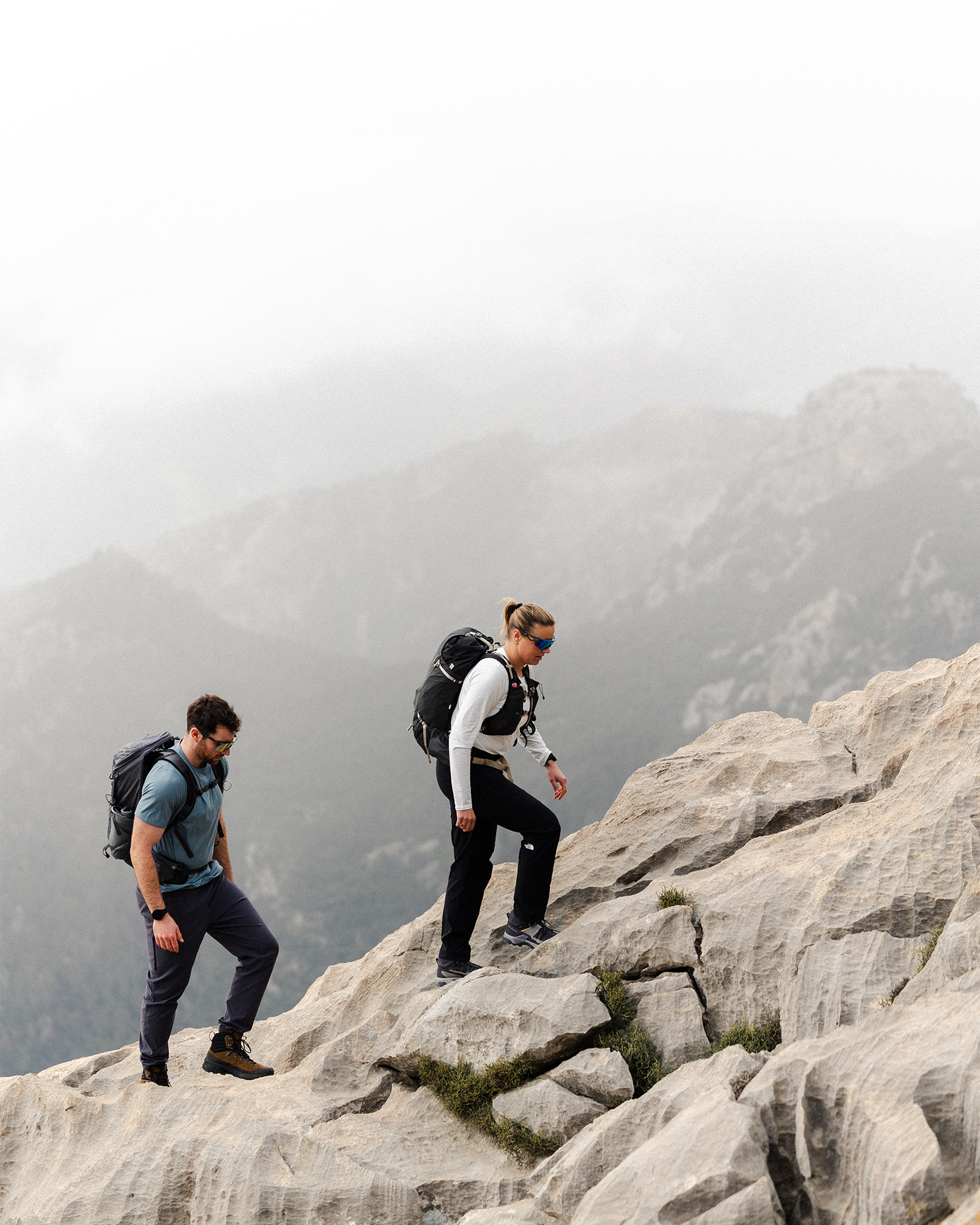 Two hikers with backpacks climb rocky terrain against a backdrop of misty mountains.
