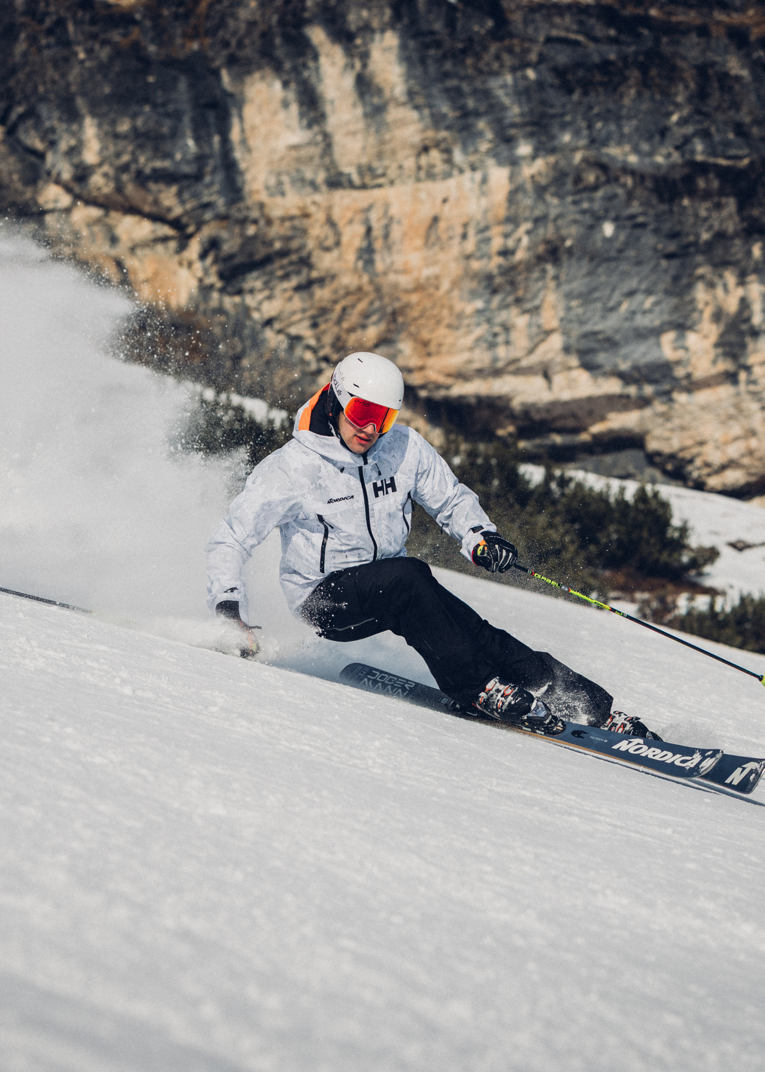 Skier in white jacket and helmet carving down a snowy slope with a rocky cliff in the background.