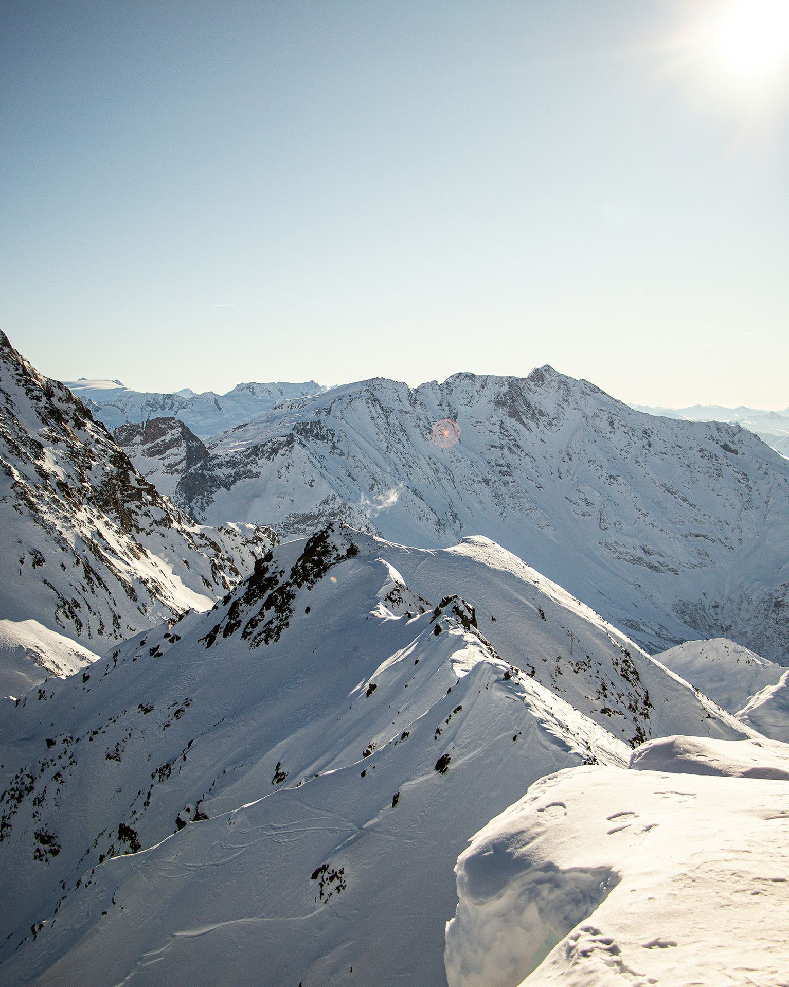 Snow-covered mountain peaks under a clear blue sky with the sun shining brightly, casting shadows on the rugged terrain.
