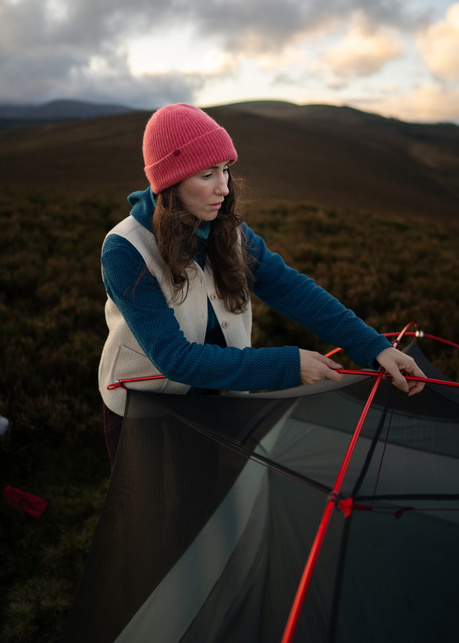 Woman in a pink beanie setting up a tent on a moorland, with rolling hills and a cloudy sky in the background.