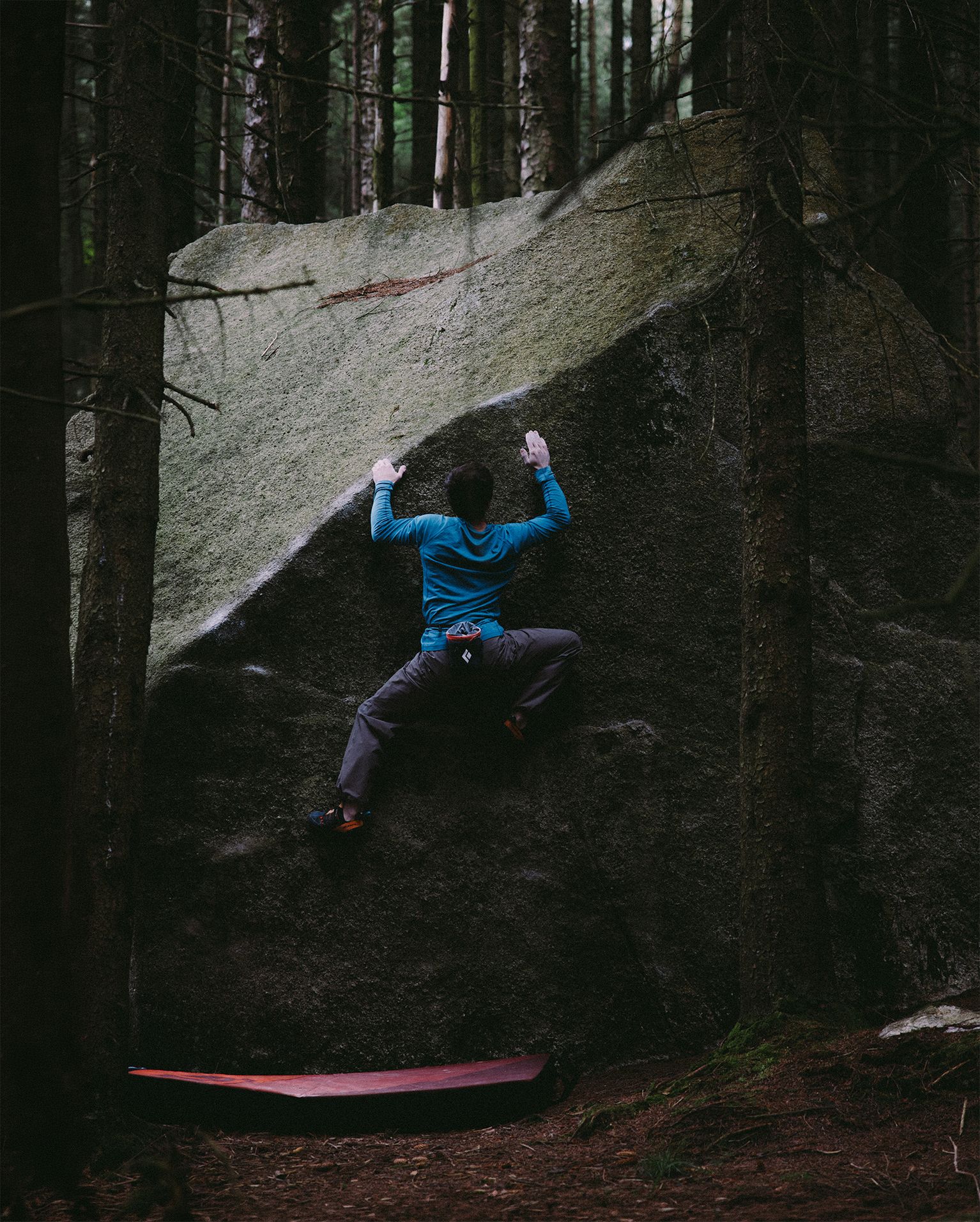 Climber wearing blue shirt and grey trousers bouldering on a large rock in a forest, with a crash pad below.