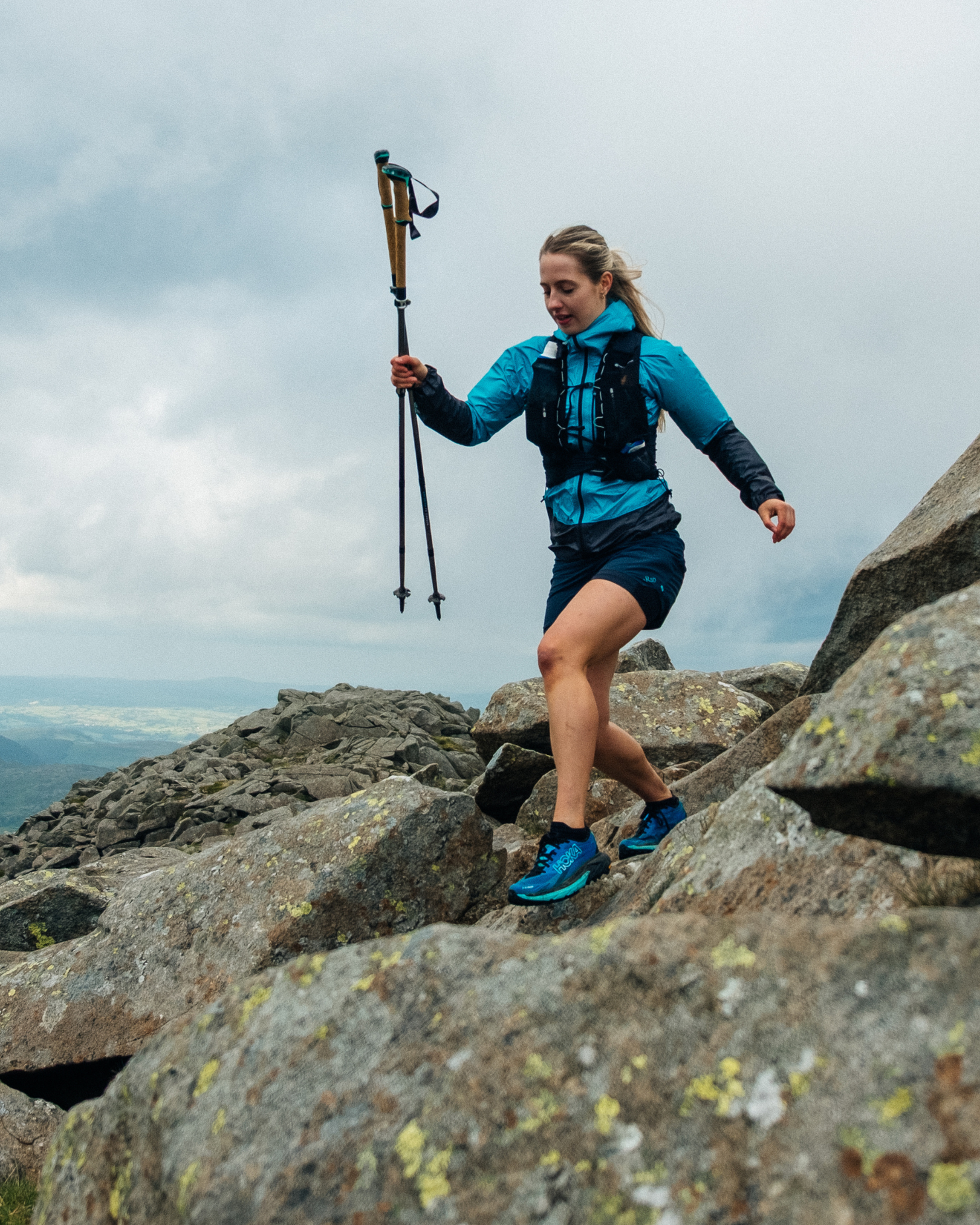 Woman in blue mountain gear descends rocky terrain with trekking poles under a cloudy sky.