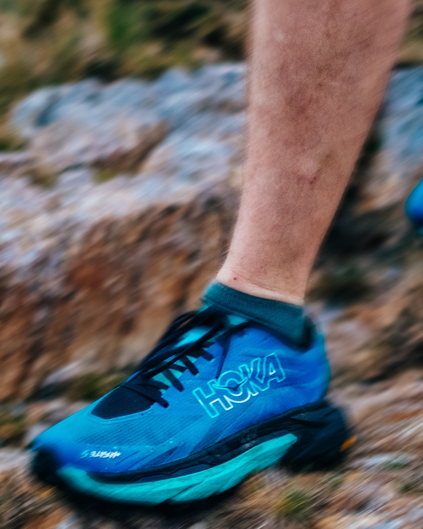 Close-up of a person wearing blue trail running shoes on rocky terrain, showcasing the shoe's design and grip.