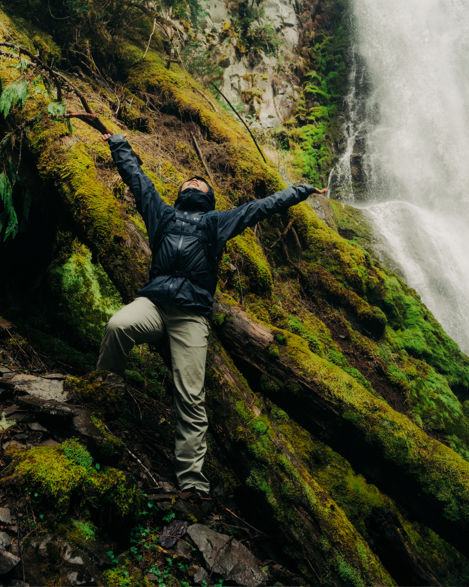 Person in outdoor gear stands on mossy rocks near a waterfall, arms spread wide, embracing the adventurous mountain environment.