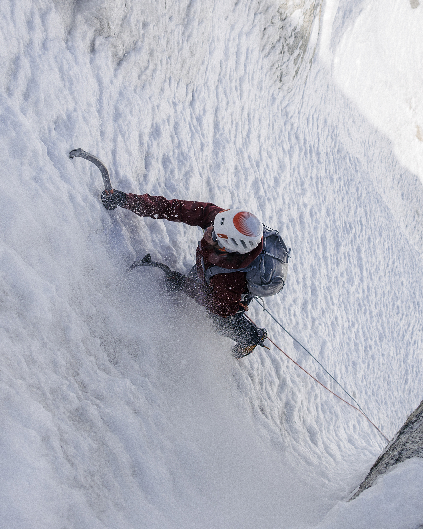 Person ice climbing a steep, snowy slope with a helmet and gear. They grip an ice axe while secured by ropes. Snow and ice surround them.