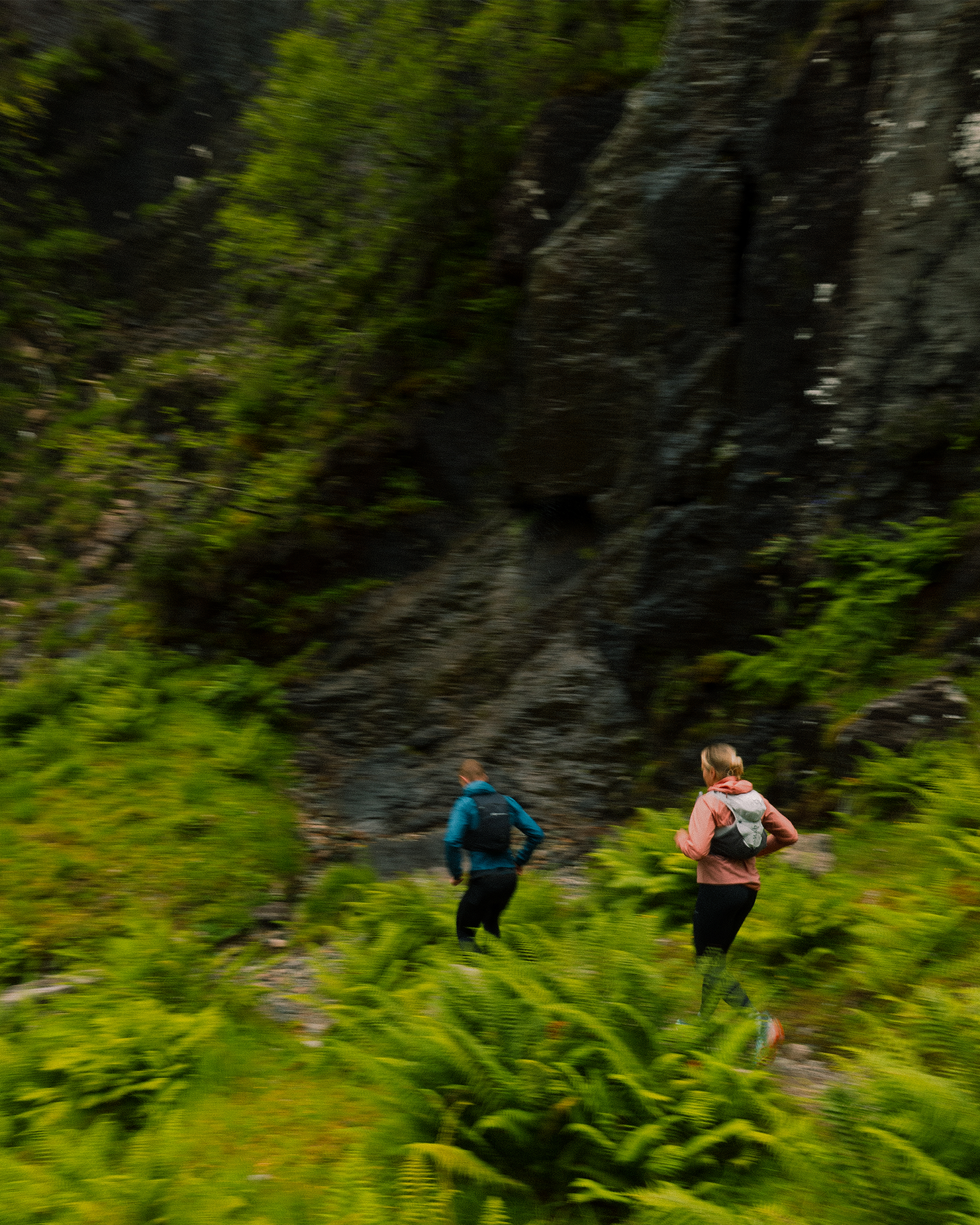 Two people with backpacks hiking through a lush, green forest with rocky terrain.