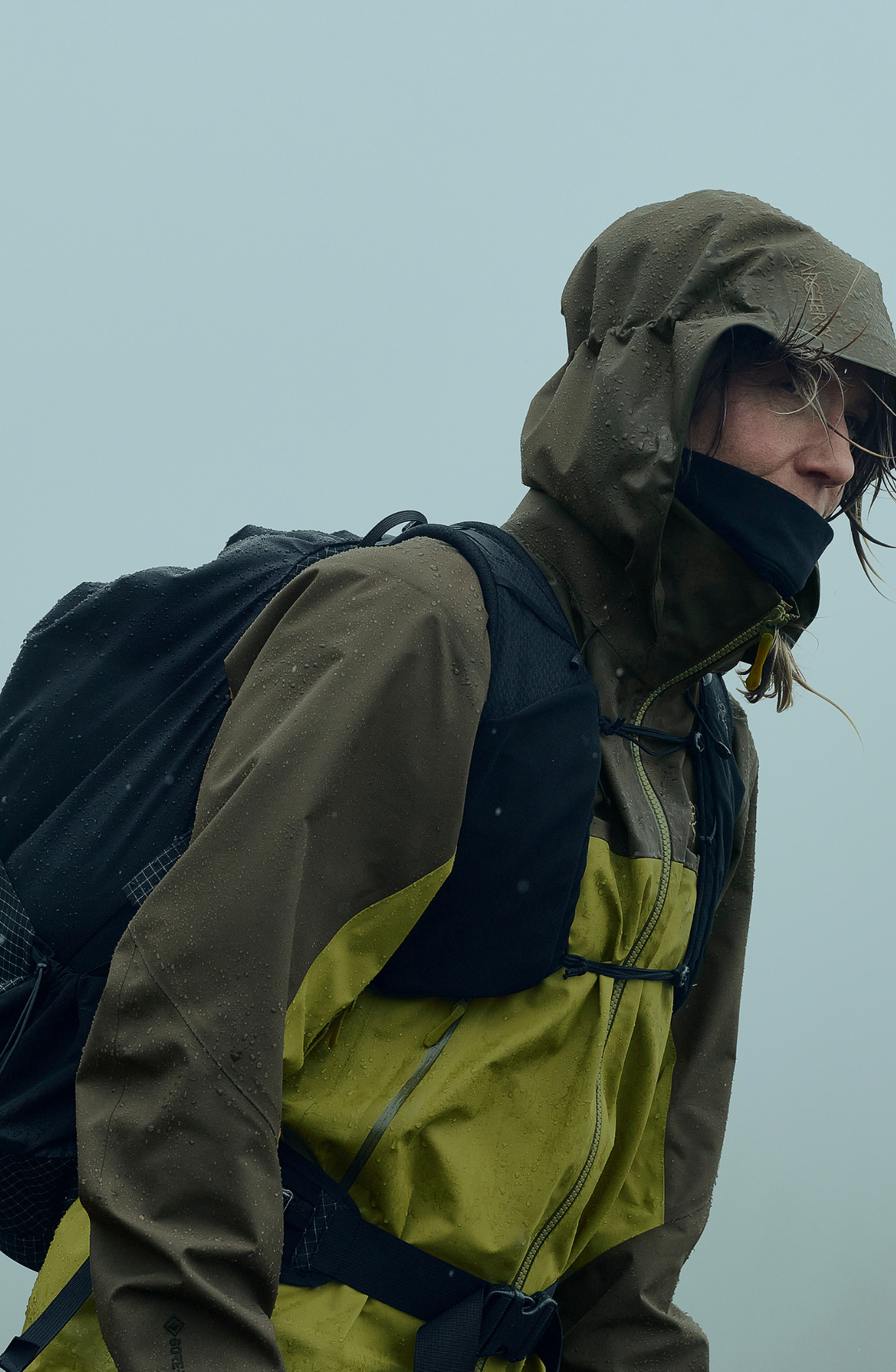 Person in green waterproof jacket and hood, carrying a black backpack, walking in misty weather.