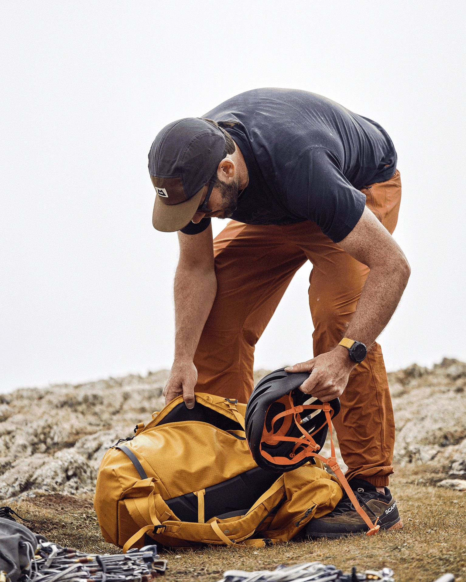 A man prepares for a climb, packing a helmet into a yellow backpack on rugged terrain, surrounded by climbing gear.
