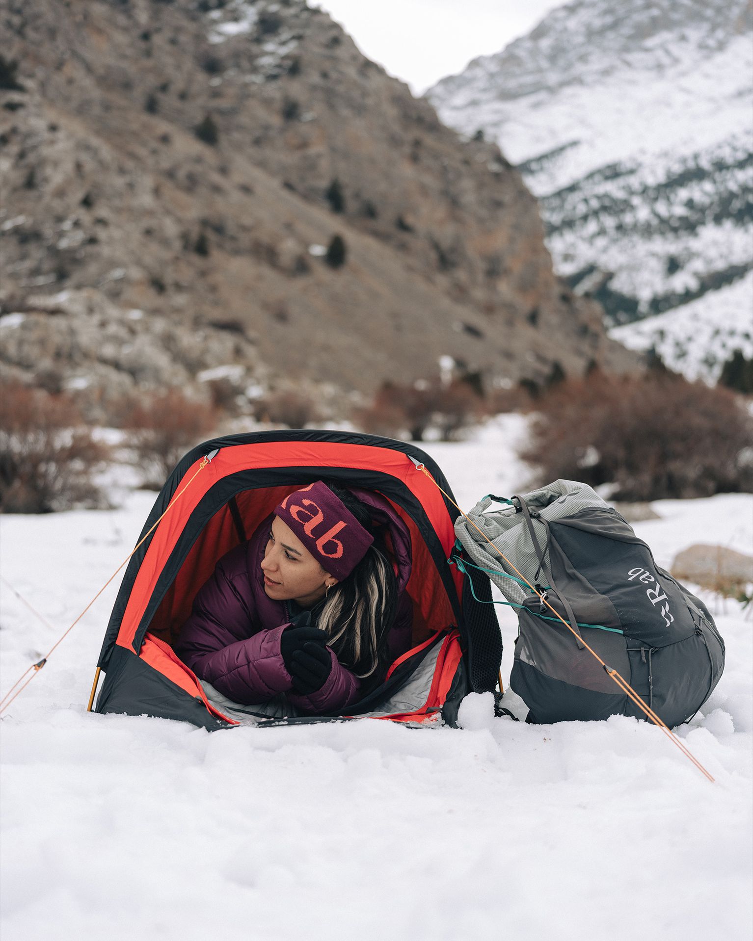 Person in a red and black bivvy on snowy ground, surrounded by mountains, with a grey backpack outside.