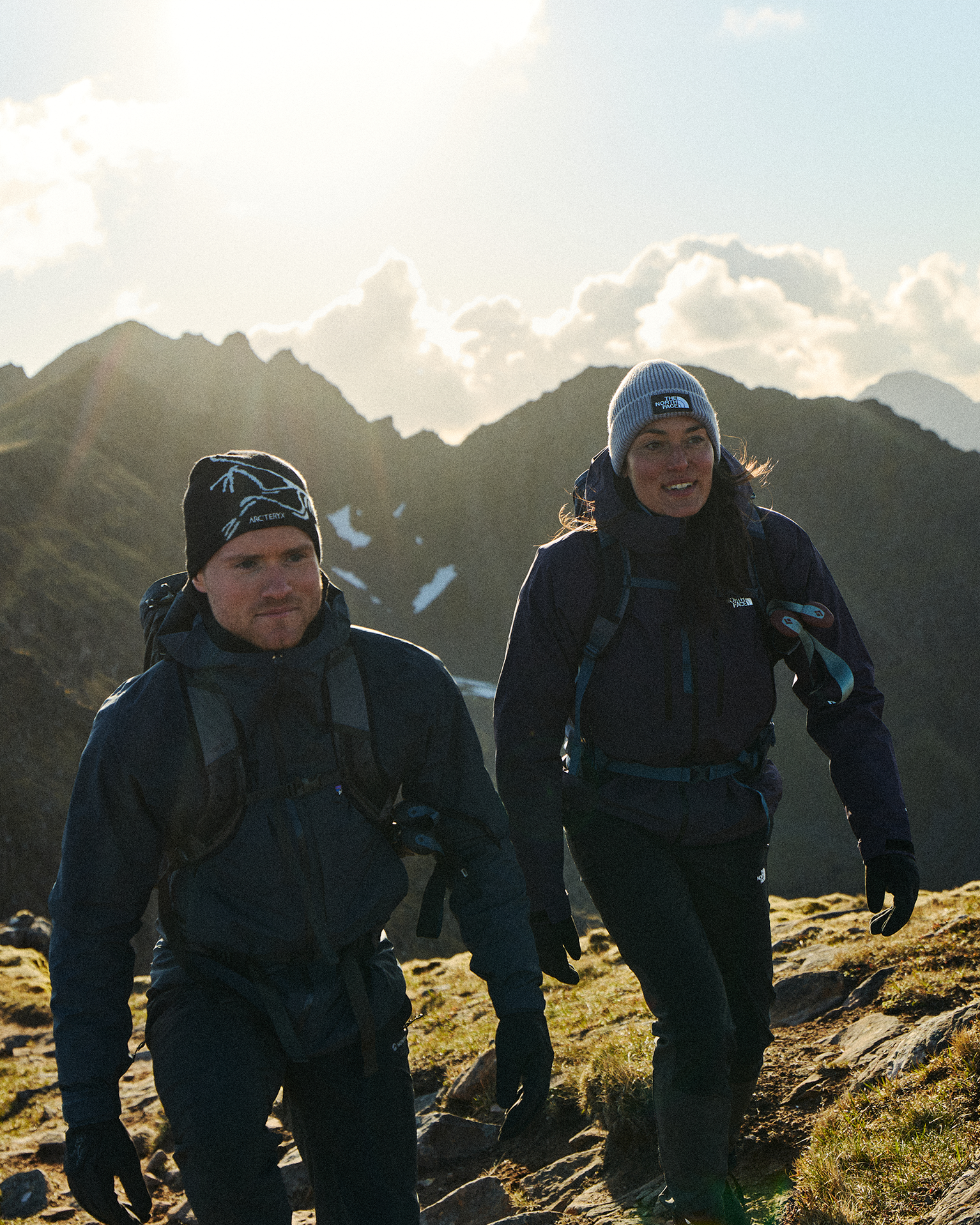 Two hikers in mountain sports gear, trekking uphill on a sunny day with rugged peaks in the background.