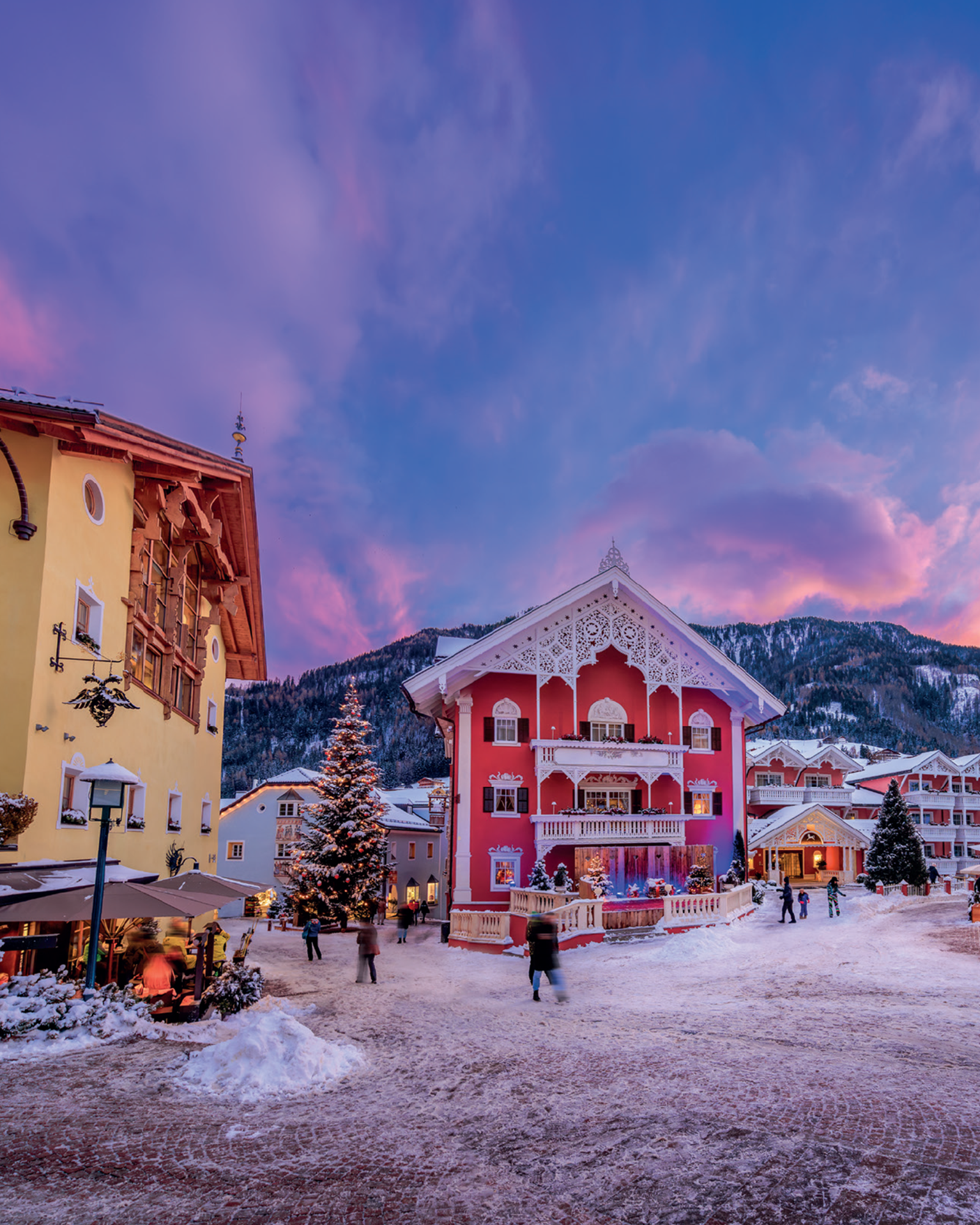 Snow-covered alpine village at twilight with colourful buildings and a decorated Christmas tree, surrounded by mountains.