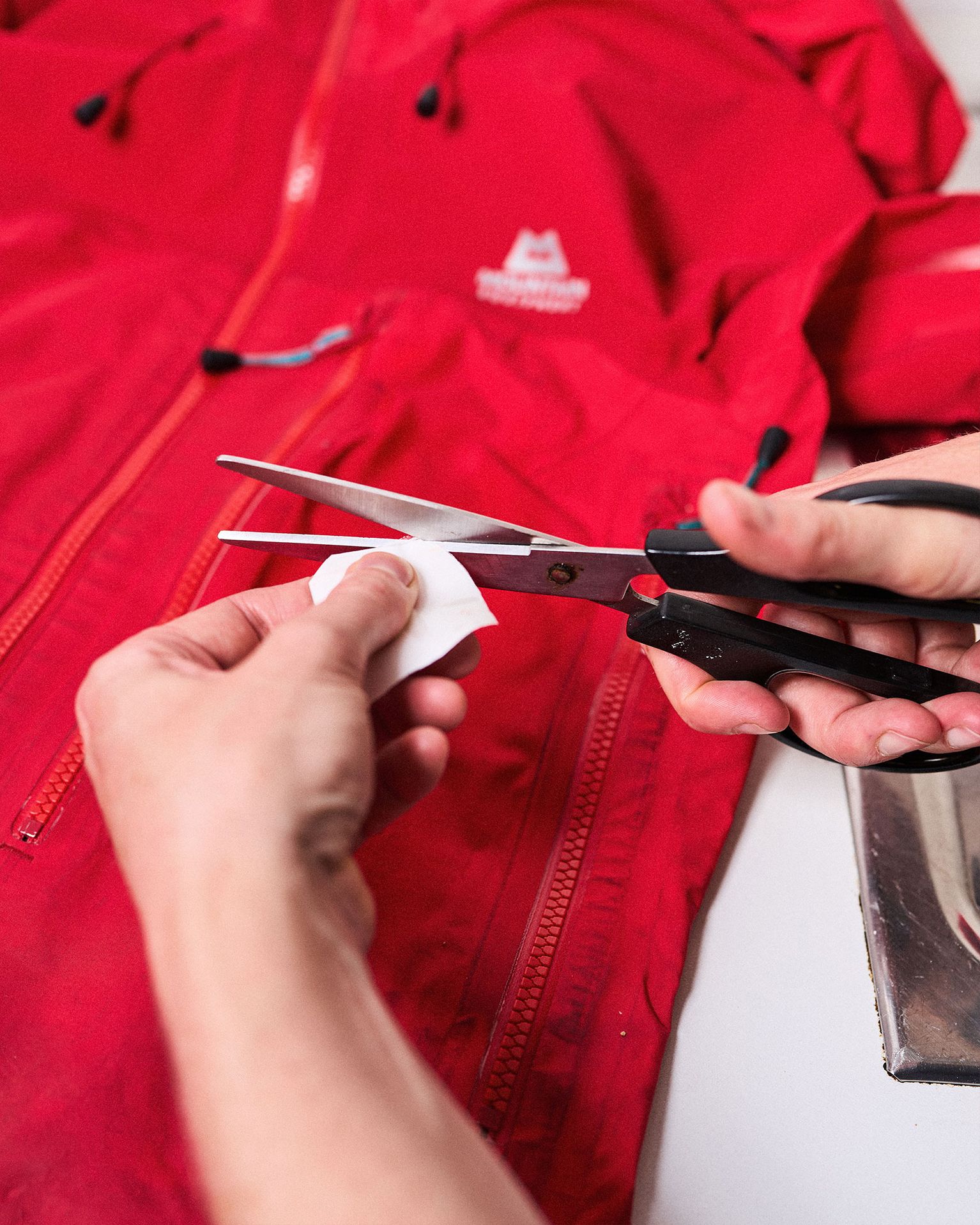 Hands cutting repair tape with scissors, preparing to repair a red jacket laid out on a table.