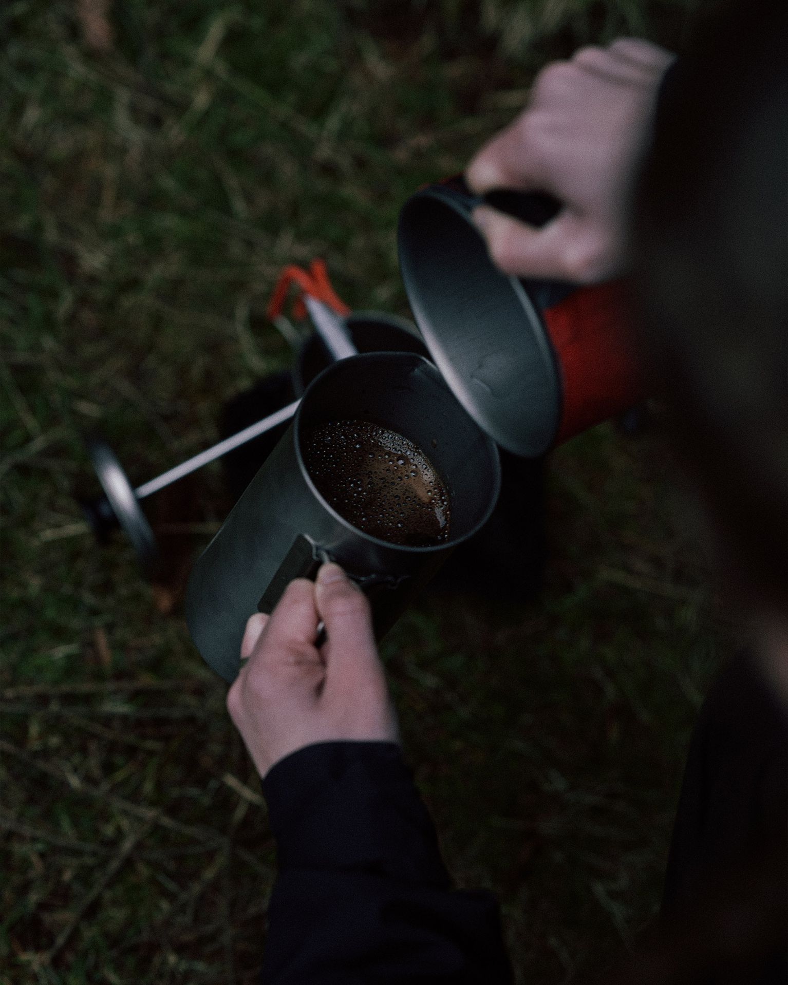 Person pouring water from a stove into a cup outdoors, with grass in the background.