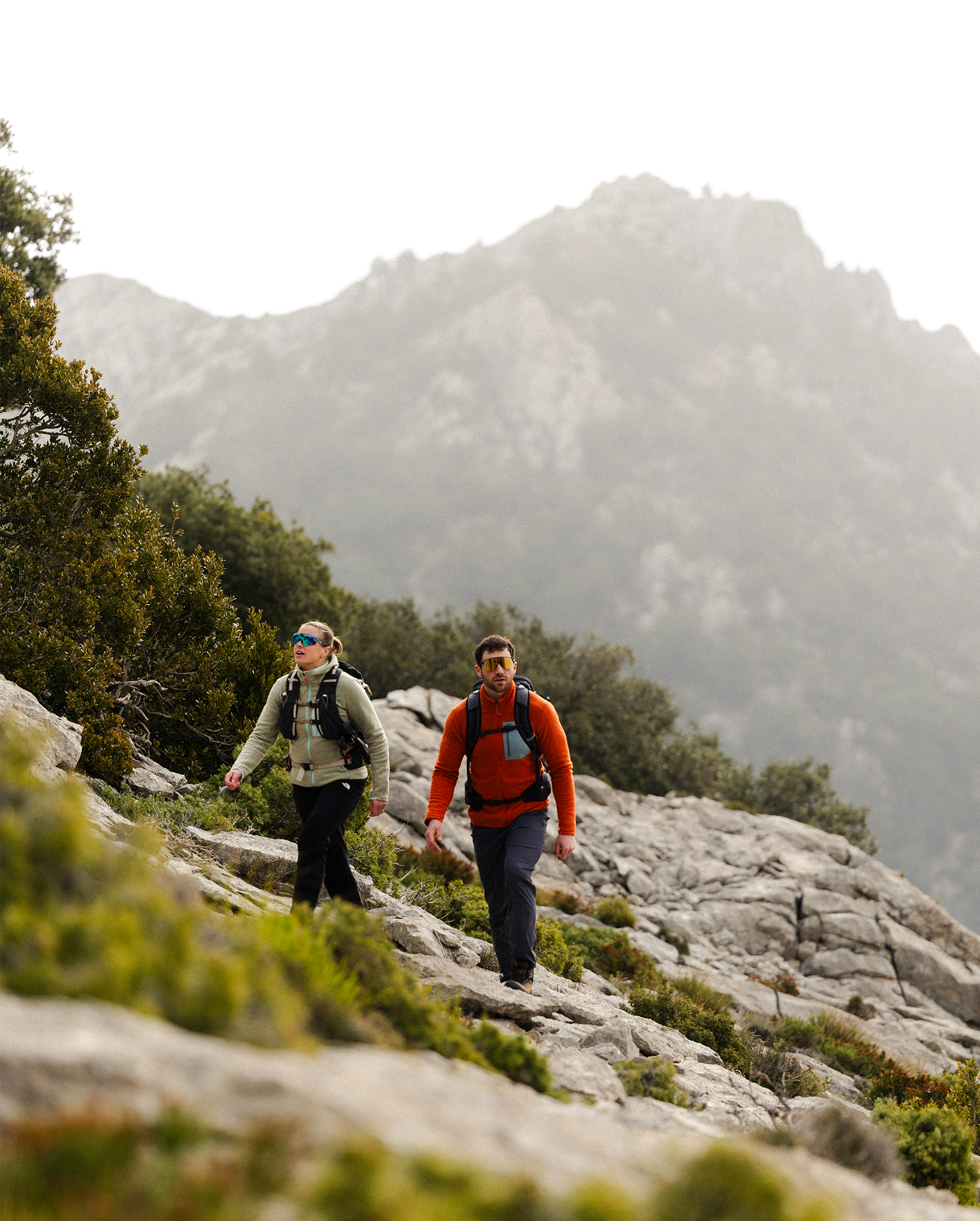Two hikers with backpacks traverse a rocky mountain slope, with a misty peak in the background.