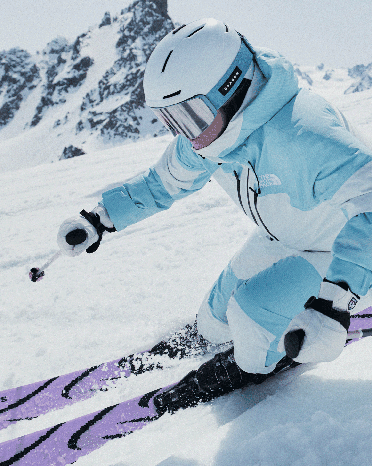 Skier in blue outfit and helmet expertly navigating snowy mountain slope with purple skis, against a backdrop of rugged peaks.