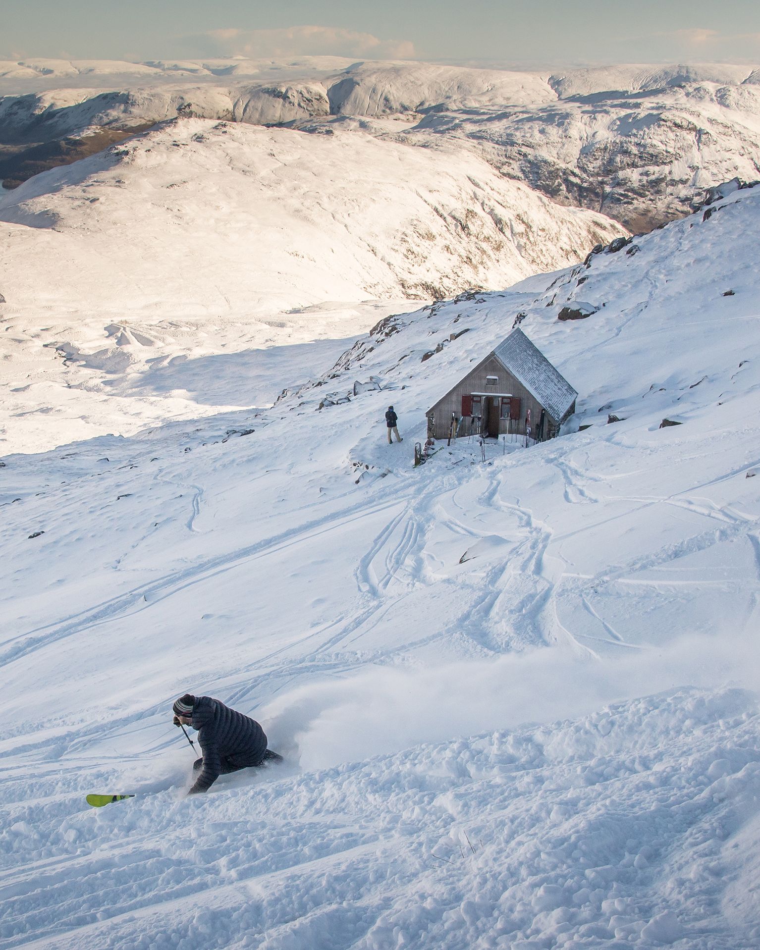Skier carving through fresh snow on a mountain slope near a small cabin, with snow-covered peaks in the background.