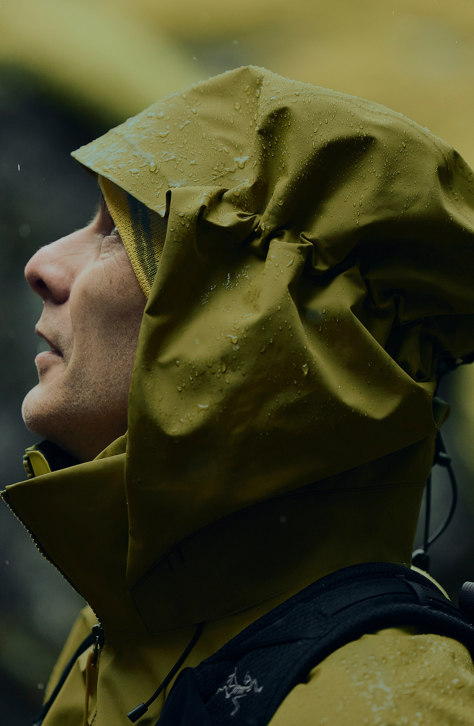 Person wearing a waterproof, olive-green jacket with hood, looking up. Raindrops visible on the fabric. Outdoor setting.