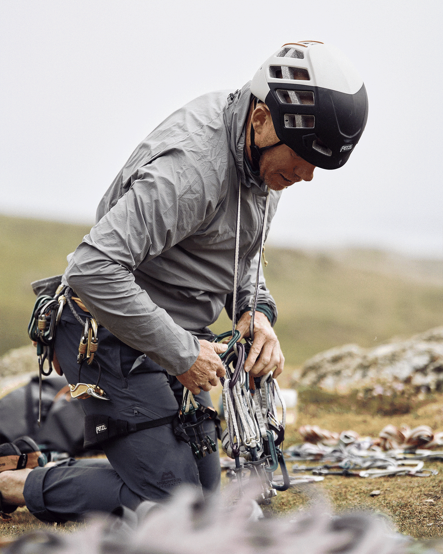 Climber in gear, kneeling on a grassy mountain, adjusting ropes and equipment, wearing a helmet and windproof jacket.