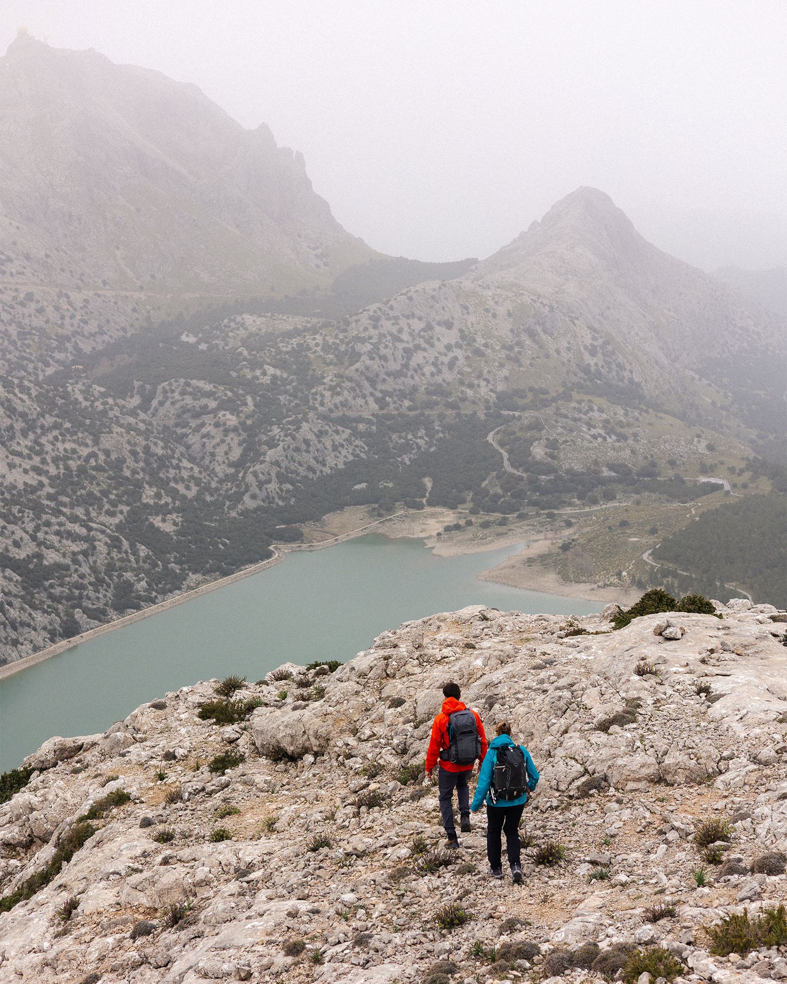 Two hikers in bright jackets trek along a rocky mountain path overlooking a serene lake and misty peaks.