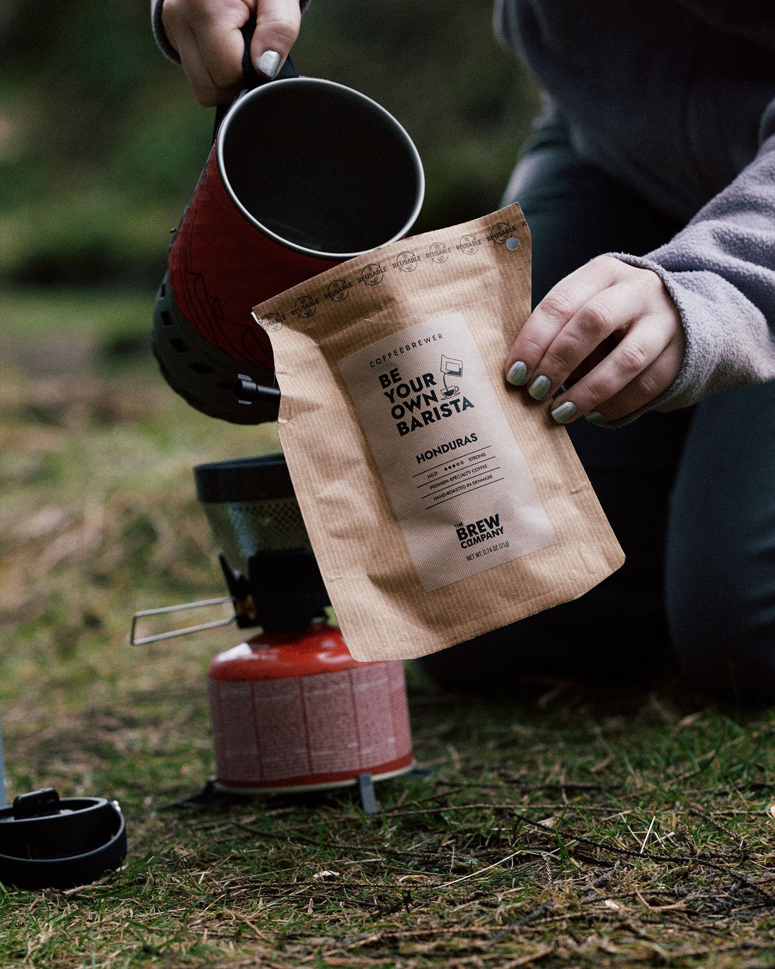 Person pouring hot water from a stove into a camping coffee bag on a portable stove outdoors.